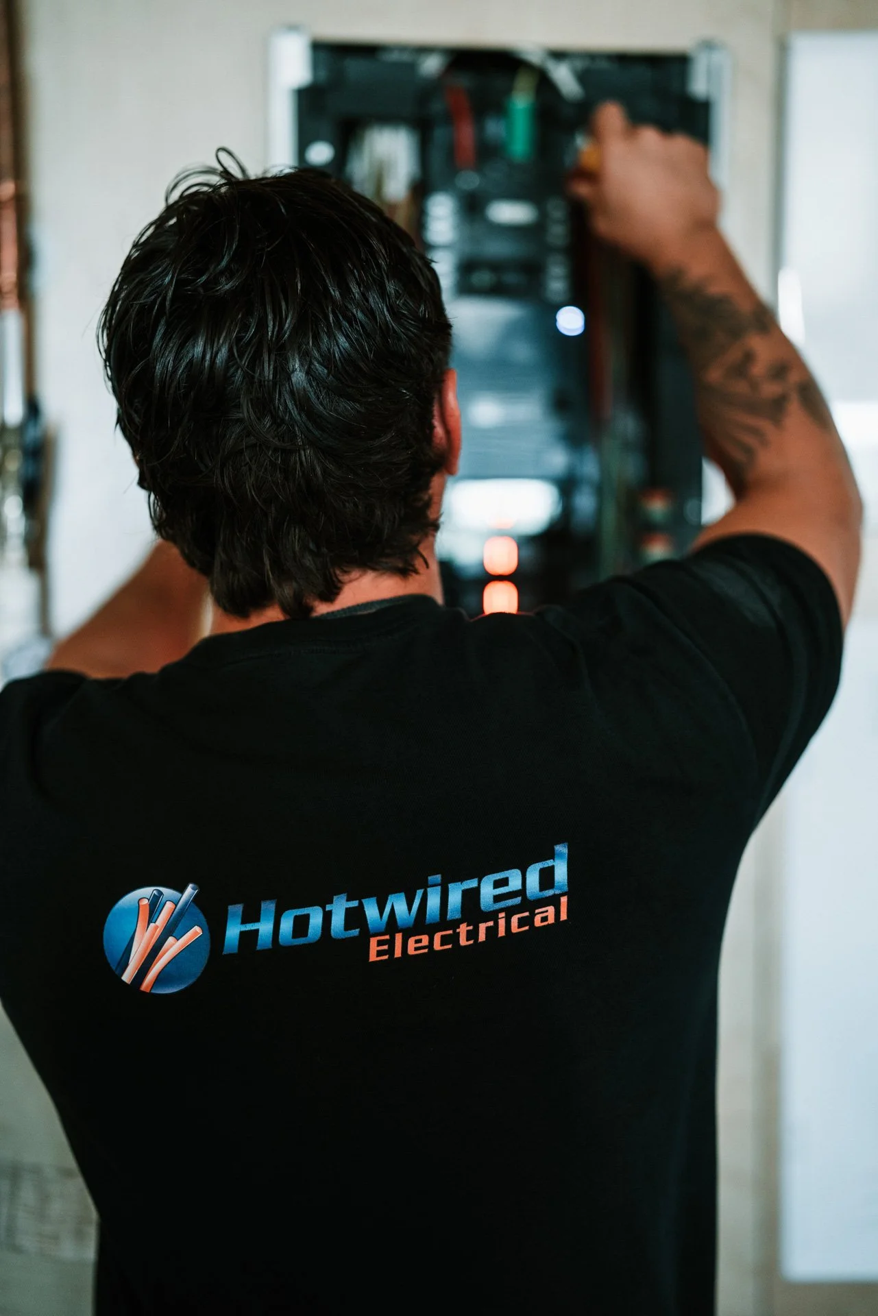 Electrician wearing a black t-shirt with 'Hotwired Electrical' logo working on an electrical panel.