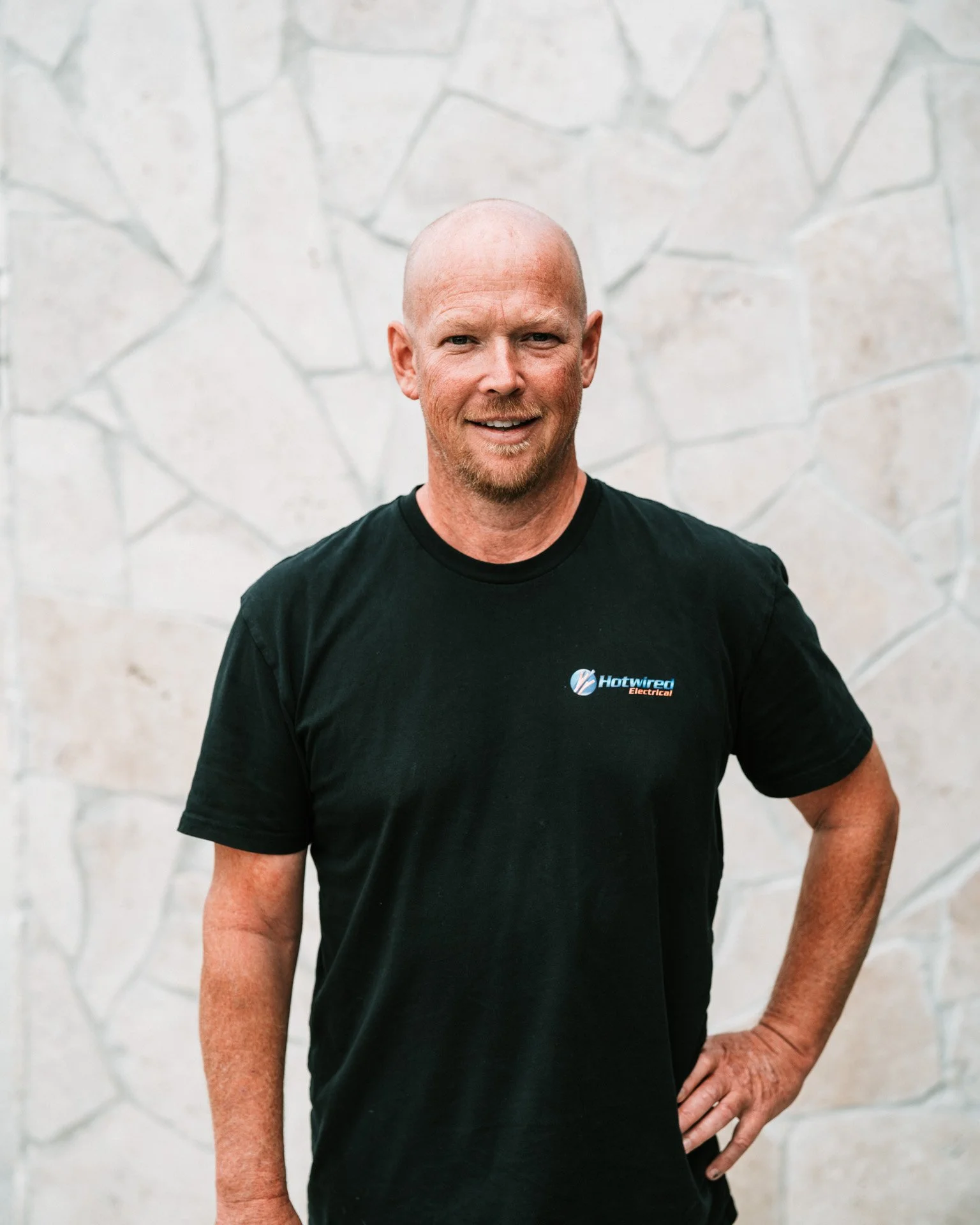 A smiling man with a bald head, light beard, wearing a black t-shirt with the logo 'Hotwired Electrical', standing against a stone wall background.
