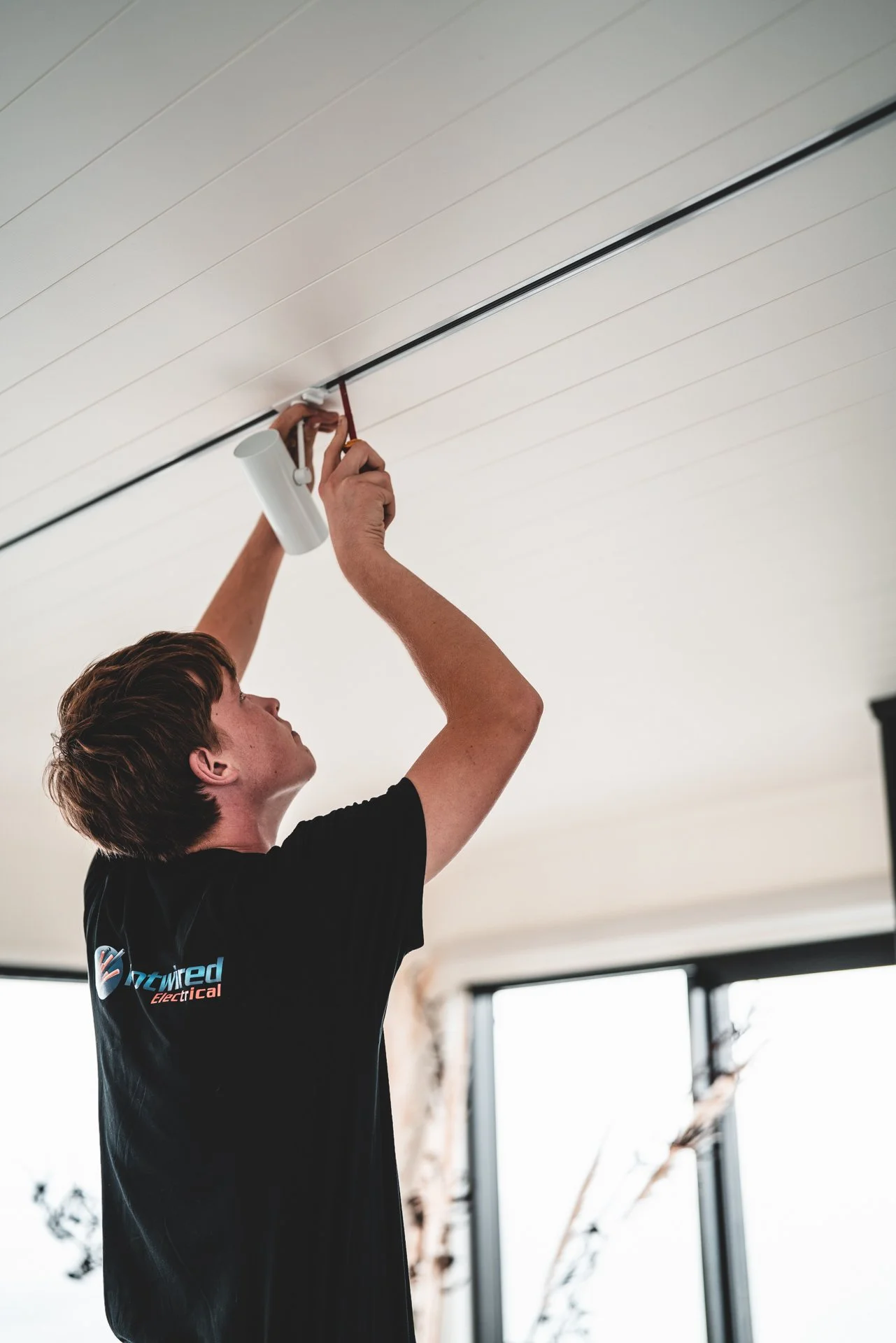A man installing a white cylindrical light fixture on a white ceiling, using a red screwdriver, inside a room with large windows.