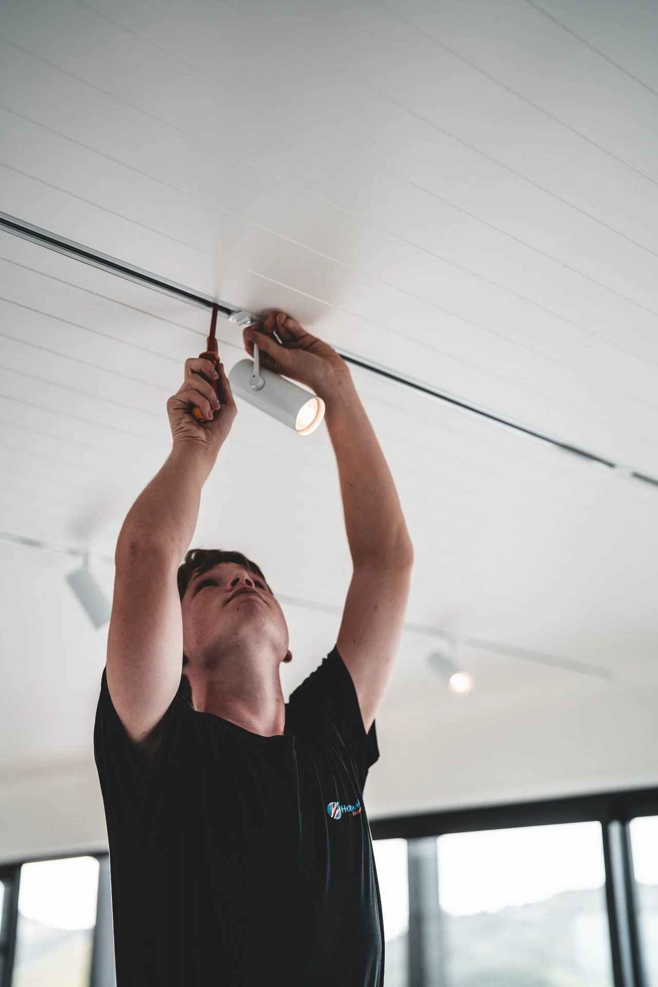 A person installing or adjusting a ceiling track light in a modern room with large windows.
