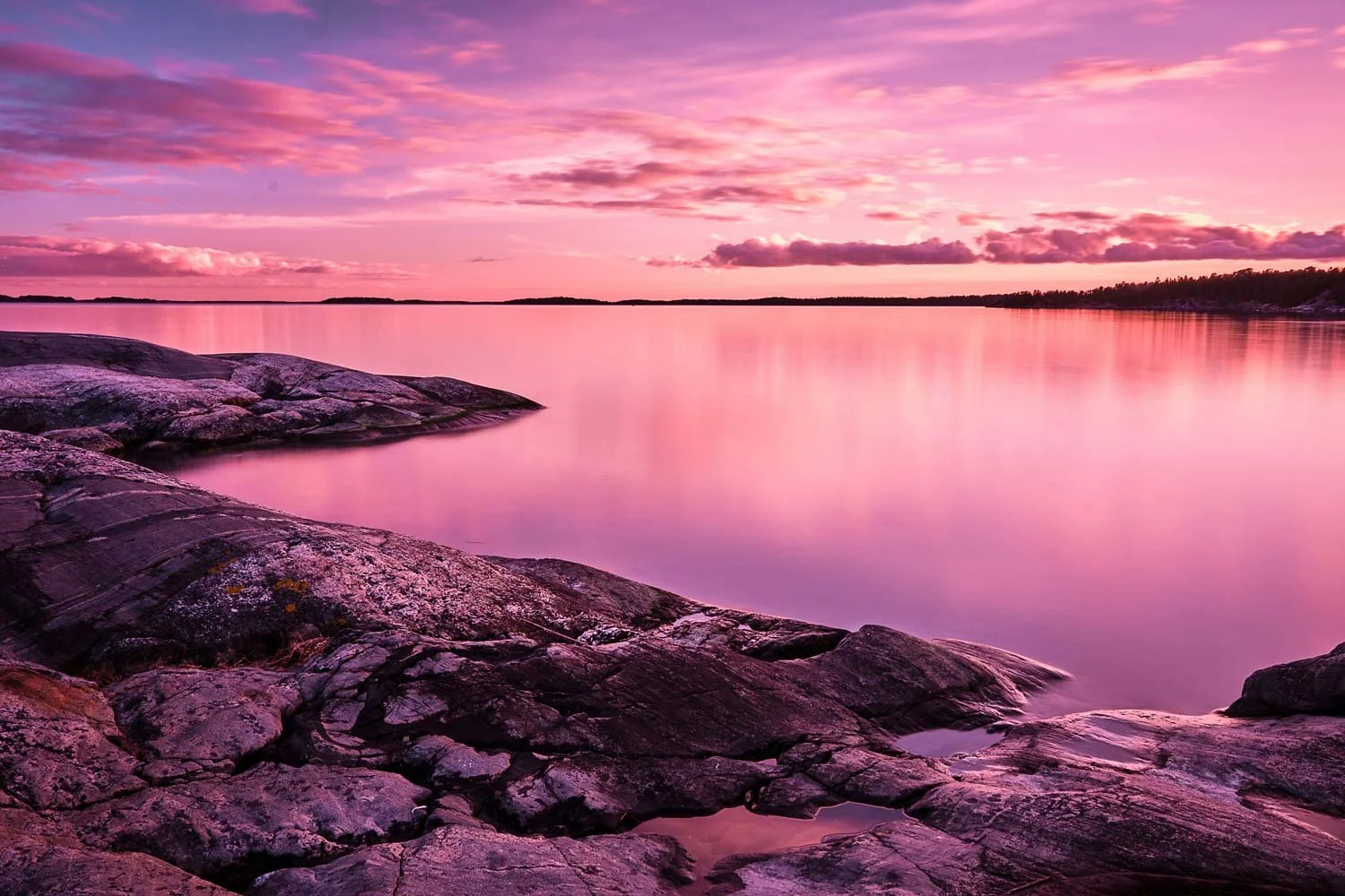 A serene scene of pink and purple clouds at sunset over a calm lake, with large rocks along the shoreline in the foreground.