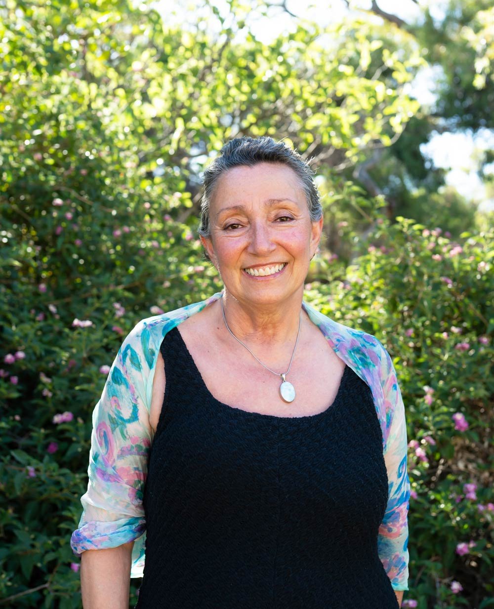 Fremantle Hypnosis Clinic founder Christina Manfredi, standing outdoors among green bushes and trees, wearing a black top and a colorful, partially sheer jacket, with a silver necklace featuring a large white pendant.