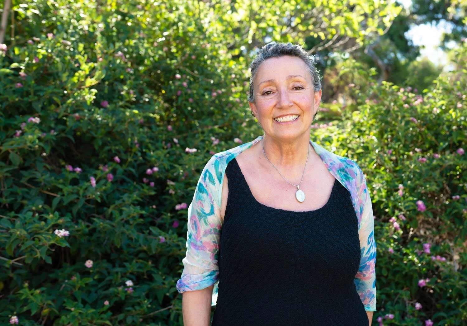 Fremantle Hypnosis Clinic founder Christina Manfredi, standing outdoors in front of green bushes with small pink flowers, wearing a black dress with a colorful sheer shawl and a pendant necklace.