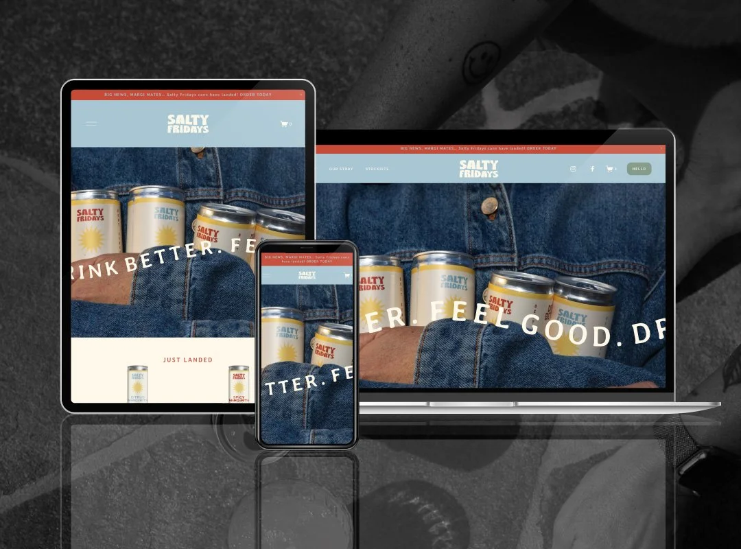 Multiple electronic devices displaying a website for Salty Fridays, featuring cans of a beverage, with a denim fabric background and a black and white surface underneath.