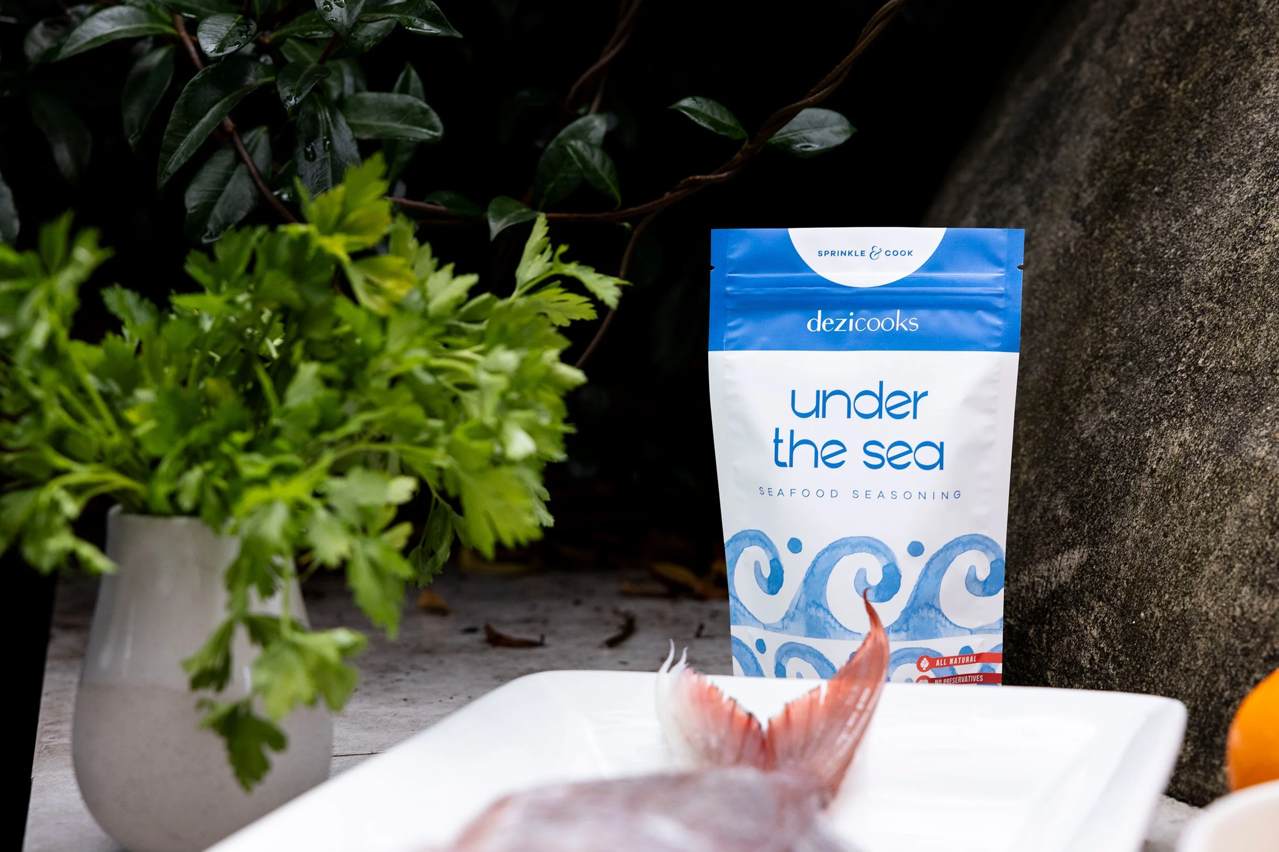 A packet of 'under the sea' seafood seasoning placed on a surface with some fresh green herbs nearby and a fish tail in the foreground.