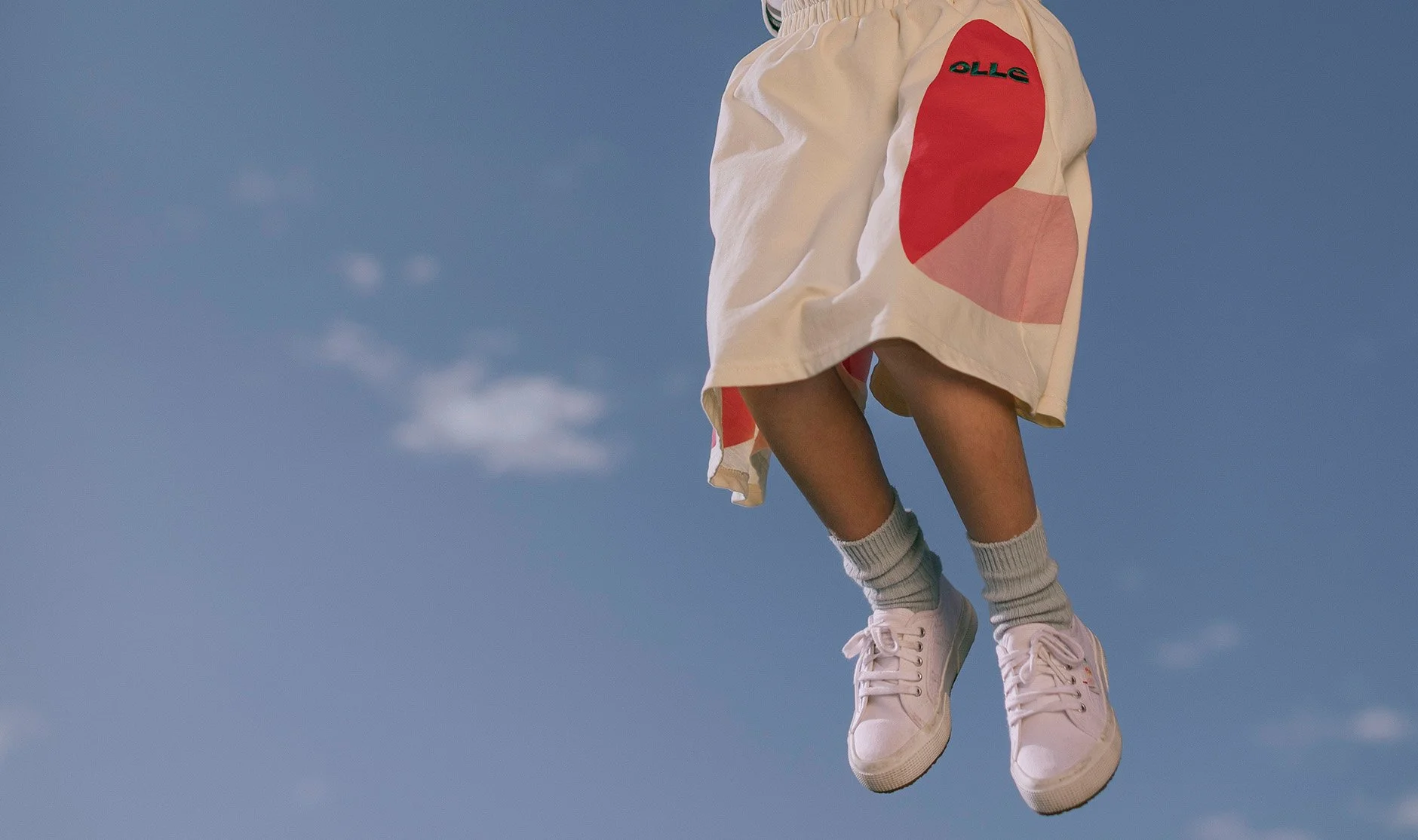 Person wearing white sneakers, gray socks, and a white skirt with red and pink circular patterns, floating against a clear blue sky.