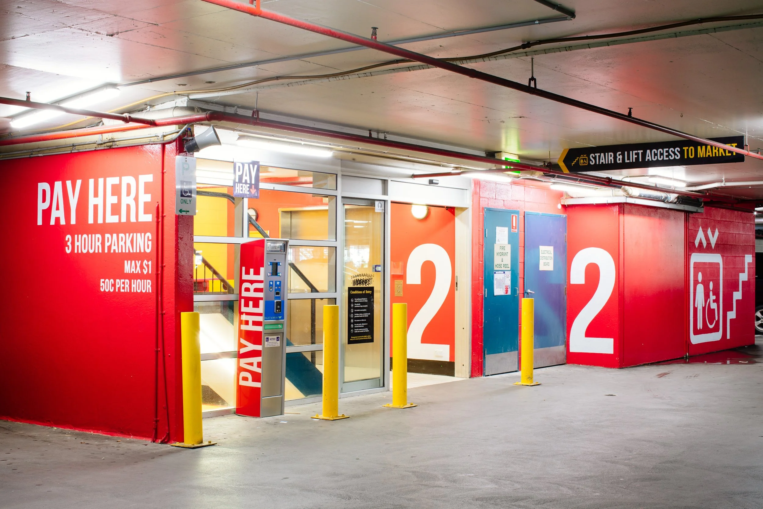 Multi-level parking garage with orange and red walls, parking payment kiosks, and signs indicating pay here for 3-hour parking and stair access to the market.