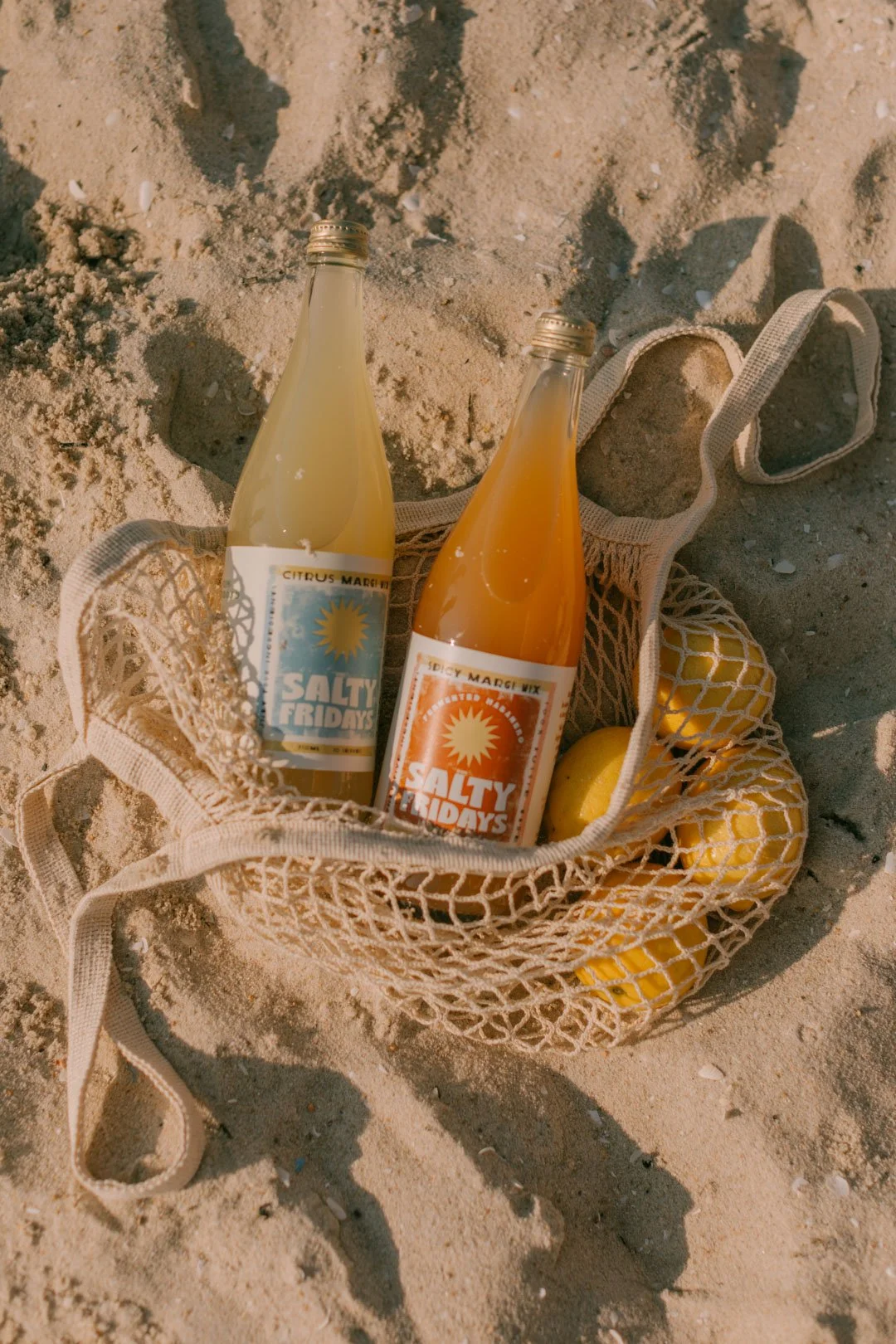 Two bottles labeled 'Salty Fridays' with citrus flavored drinks placed in a beige mesh bag on sandy beach ground, with some lemons inside the bag.