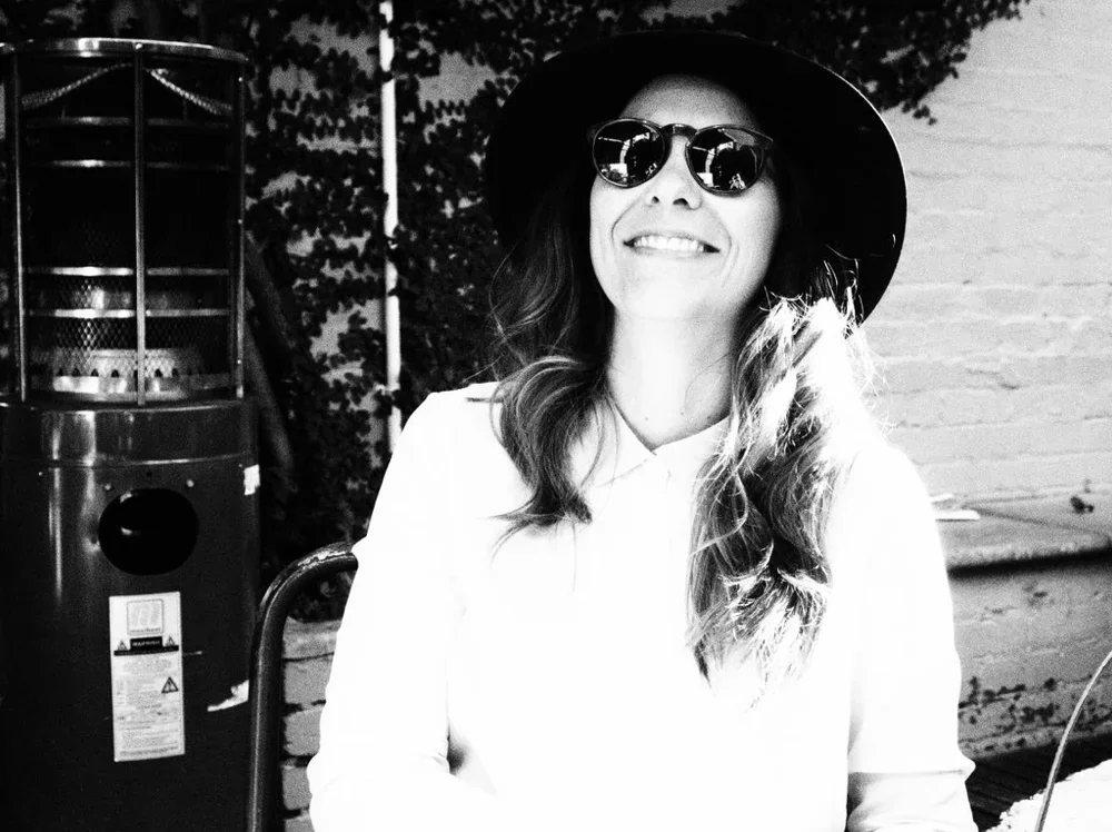 Woman wearing sunglasses and a large hat, smiling outdoors near a brick wall.