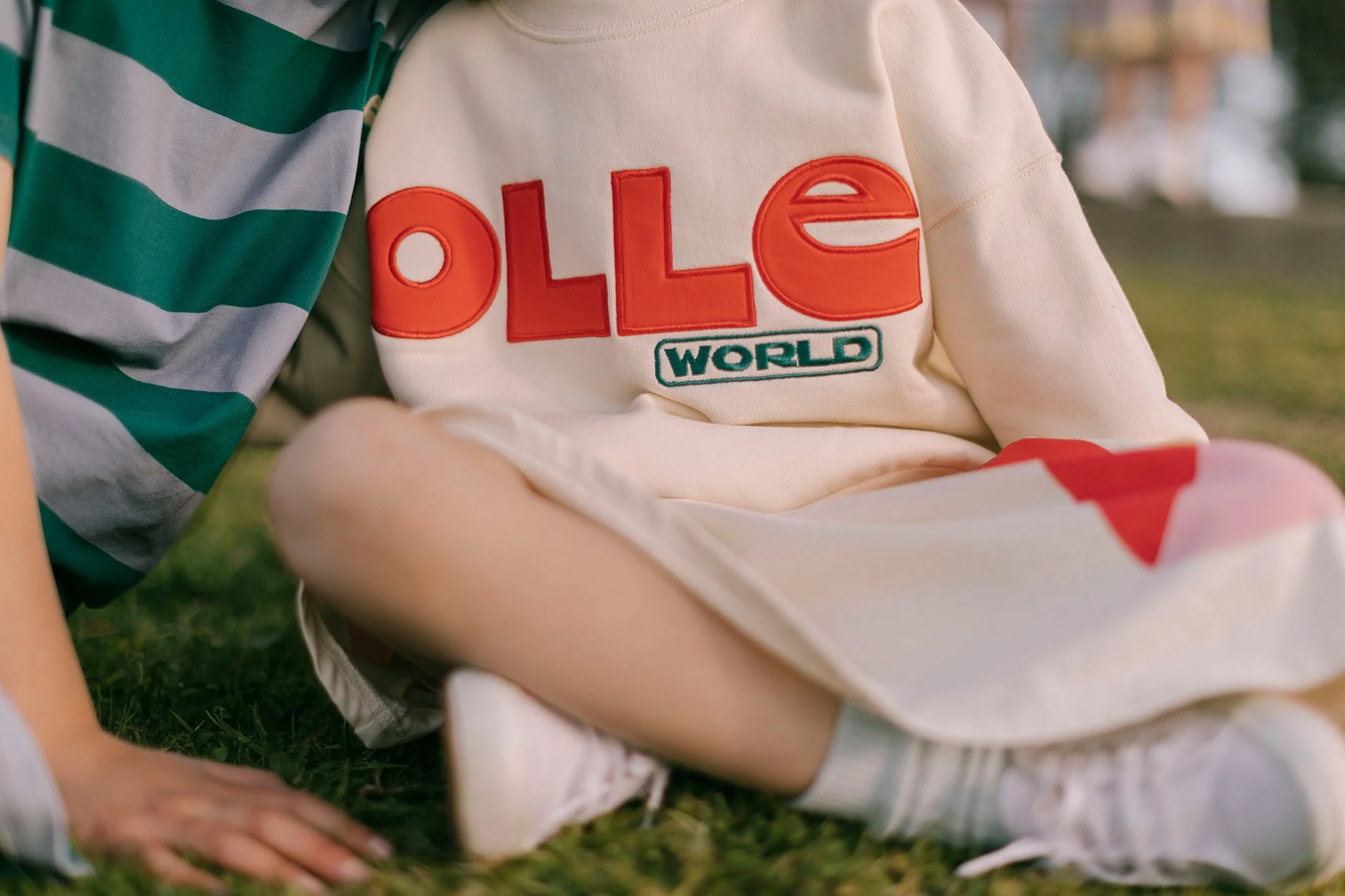 Close-up of children sitting on grass, one in a cream hoodie with 'OLLE WORLD' embroidered, and another in a green and gray striped shirt, with visible legs and hand, outdoors.