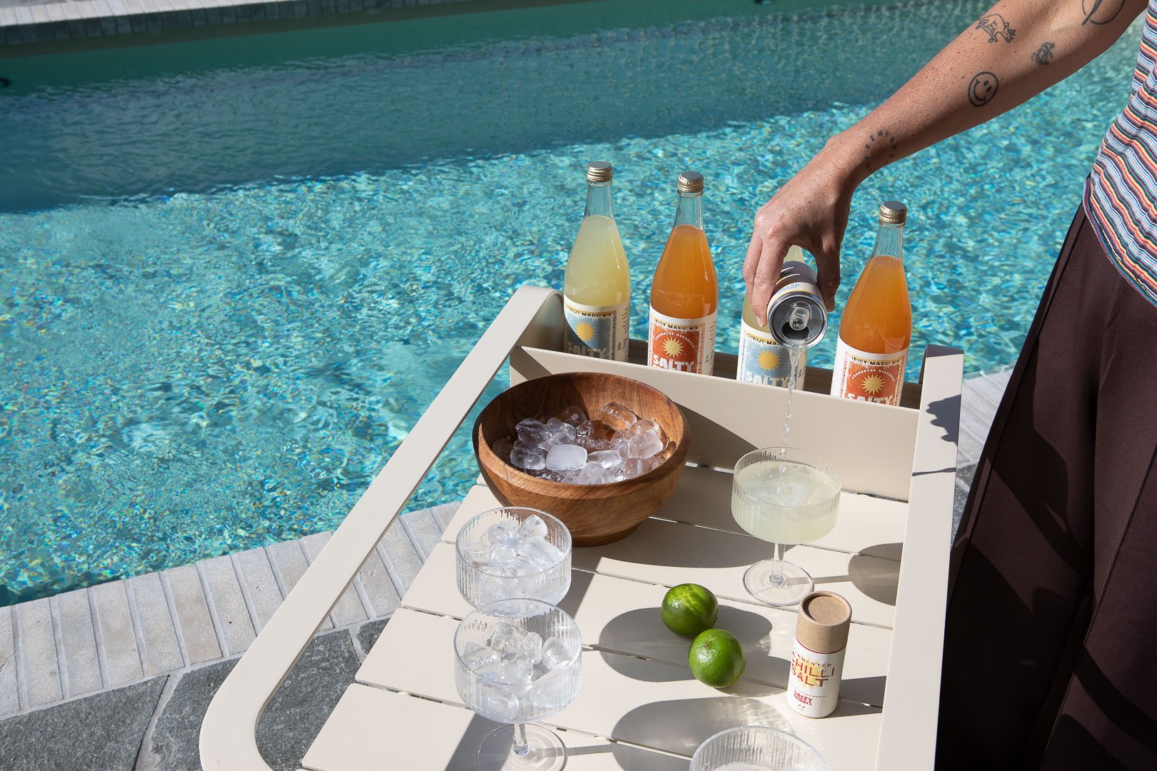 A person is pouring a drink from a can into a cocktail glass on a white poolside table. The table has three bottles of flavored mixer, a wooden bowl with ice, two glasses with ice, two limes, and a container of sea salt. The pool’s water is blue and in the background.