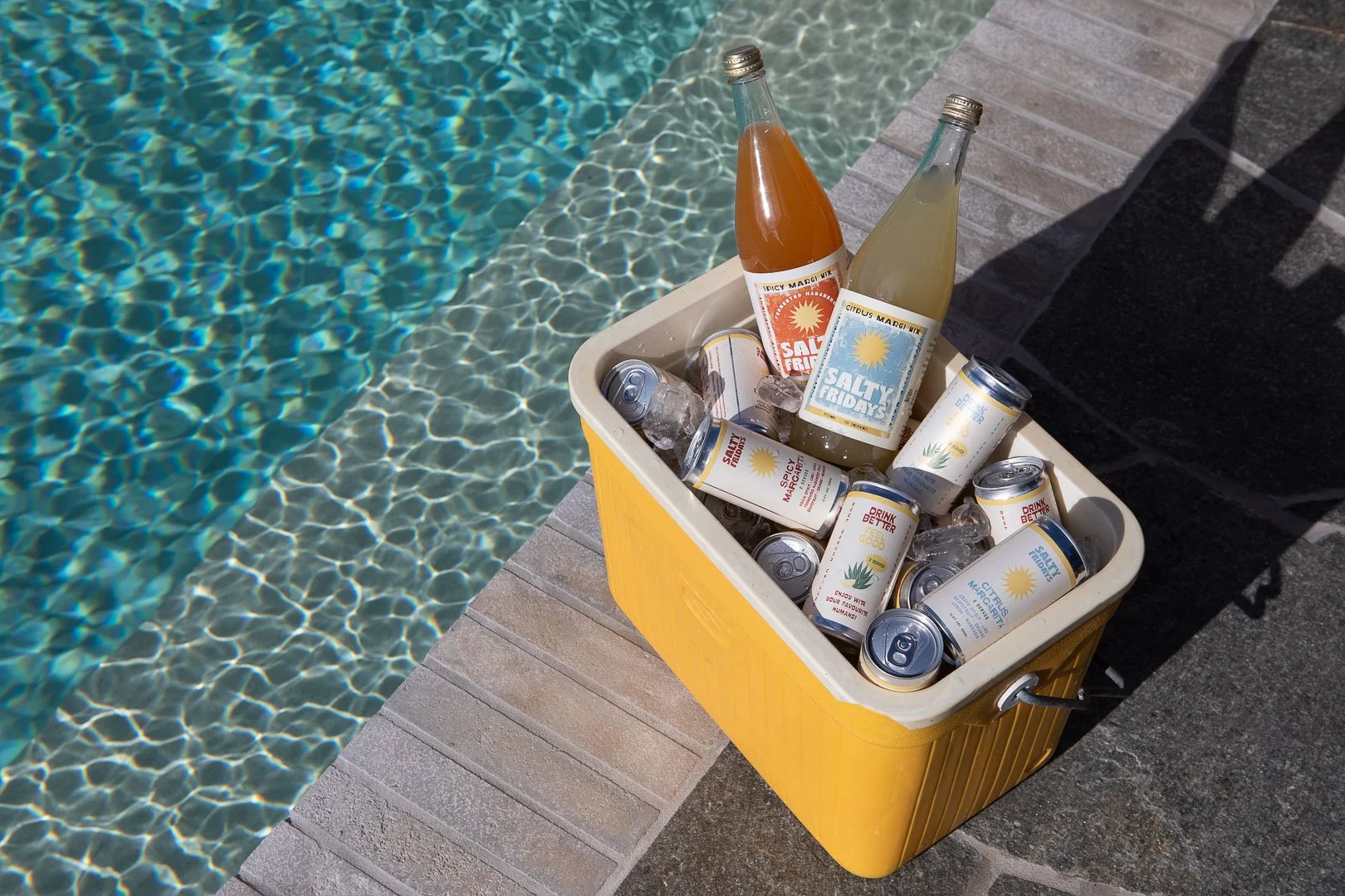 A yellow cooler filled with cans and bottles, some with colorful labels, sitting on a poolside deck with a swimming pool nearby.