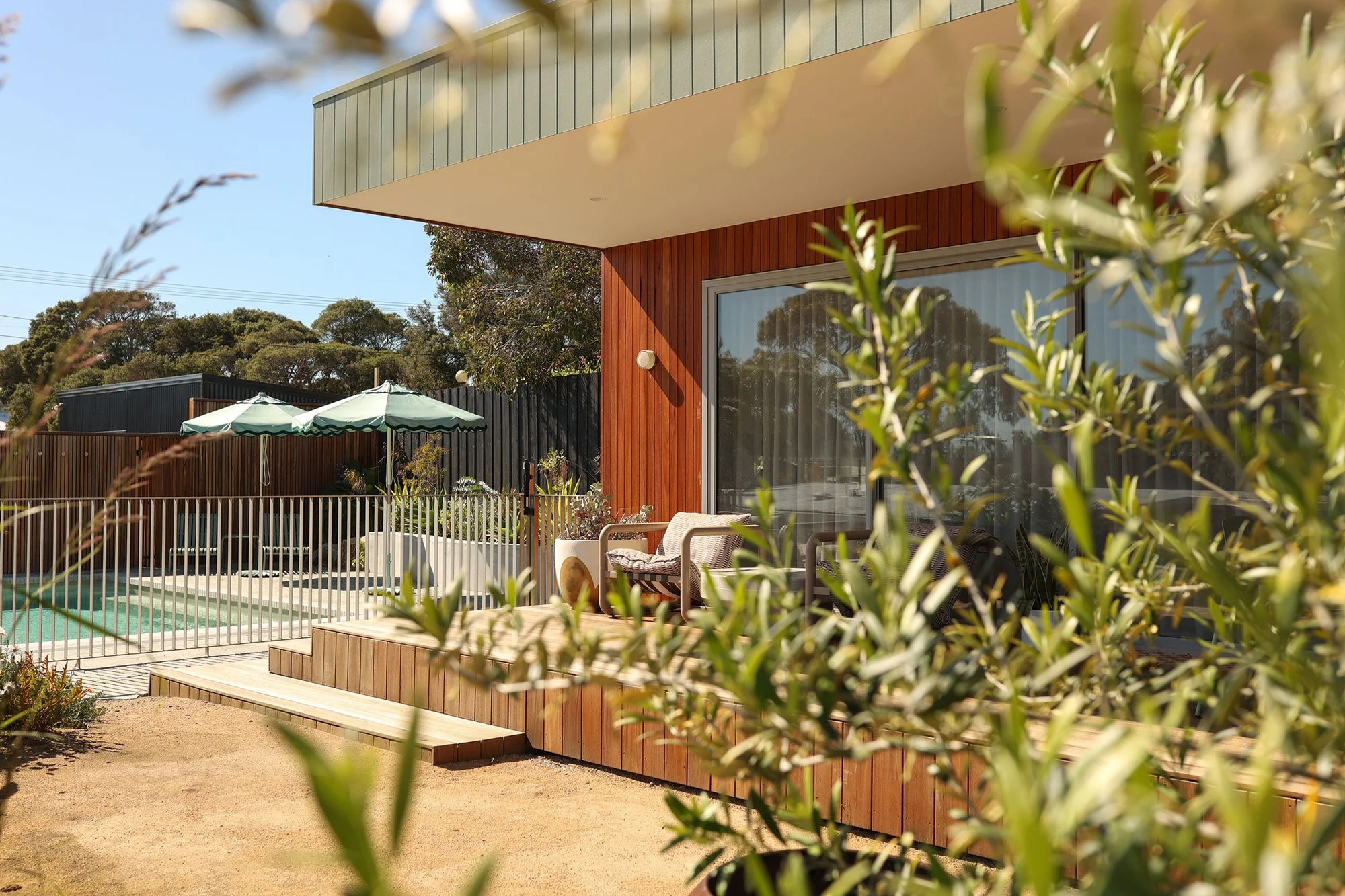 Modern house with large glass window, wooden exterior, and a balcony with outdoor furniture. The backyard has a small pool, umbrellas, and garden plants, enclosed by a black fence.