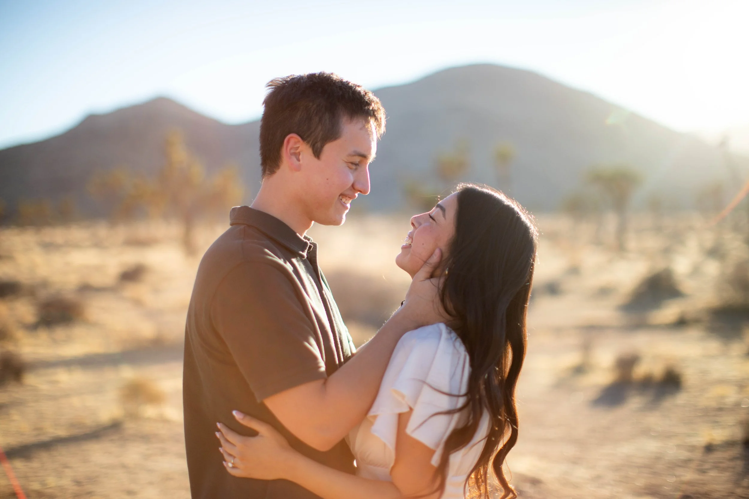 A young man and woman smiling and embracing each other outdoors in a desert landscape with mountains in the background, during sunset.