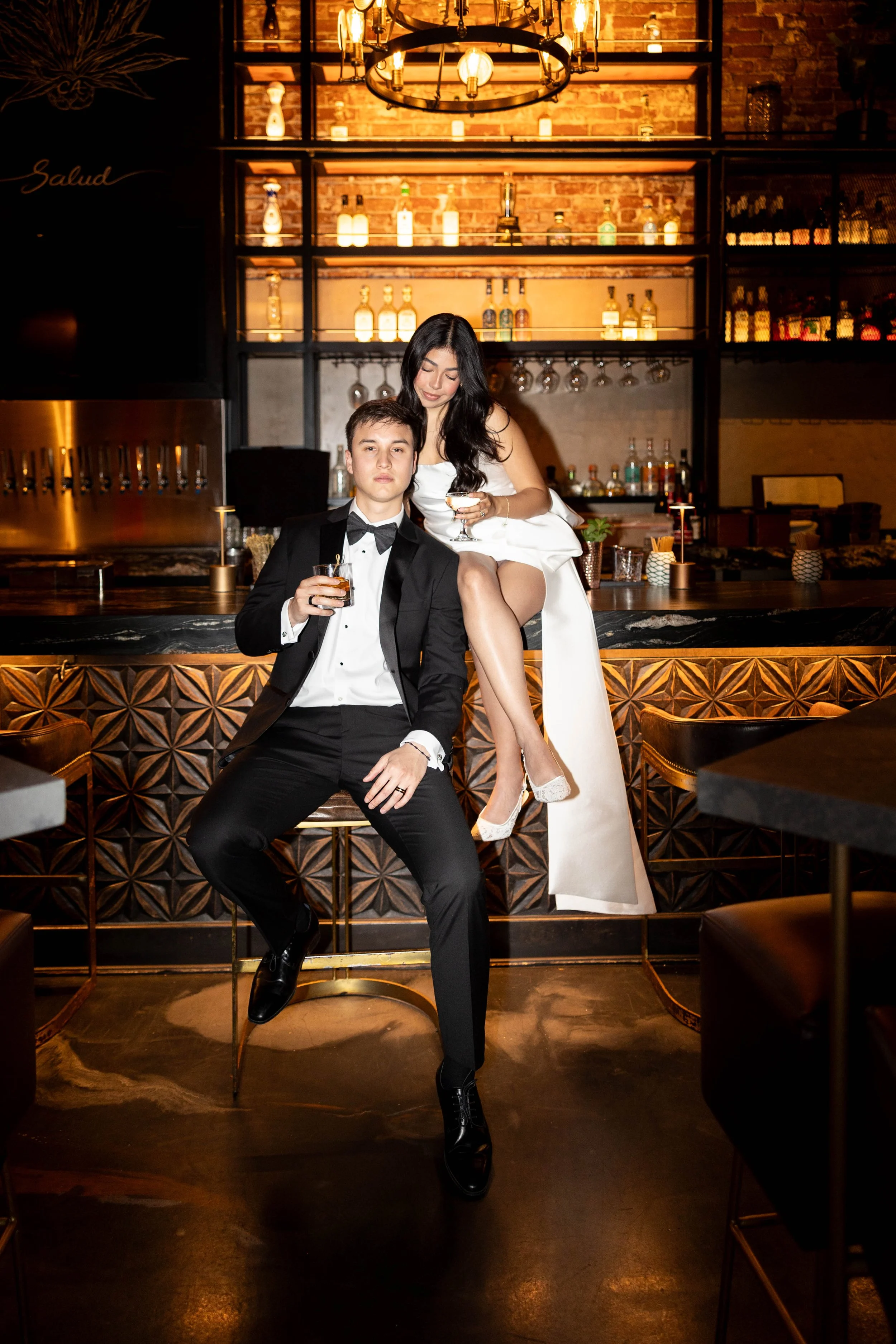 A young man in a black tuxedo and a young woman in a white dress and high heels sitting together at a bar, holding drinks, in a warmly-lit, stylish bar with liquor bottles and glasses on shelves behind them.