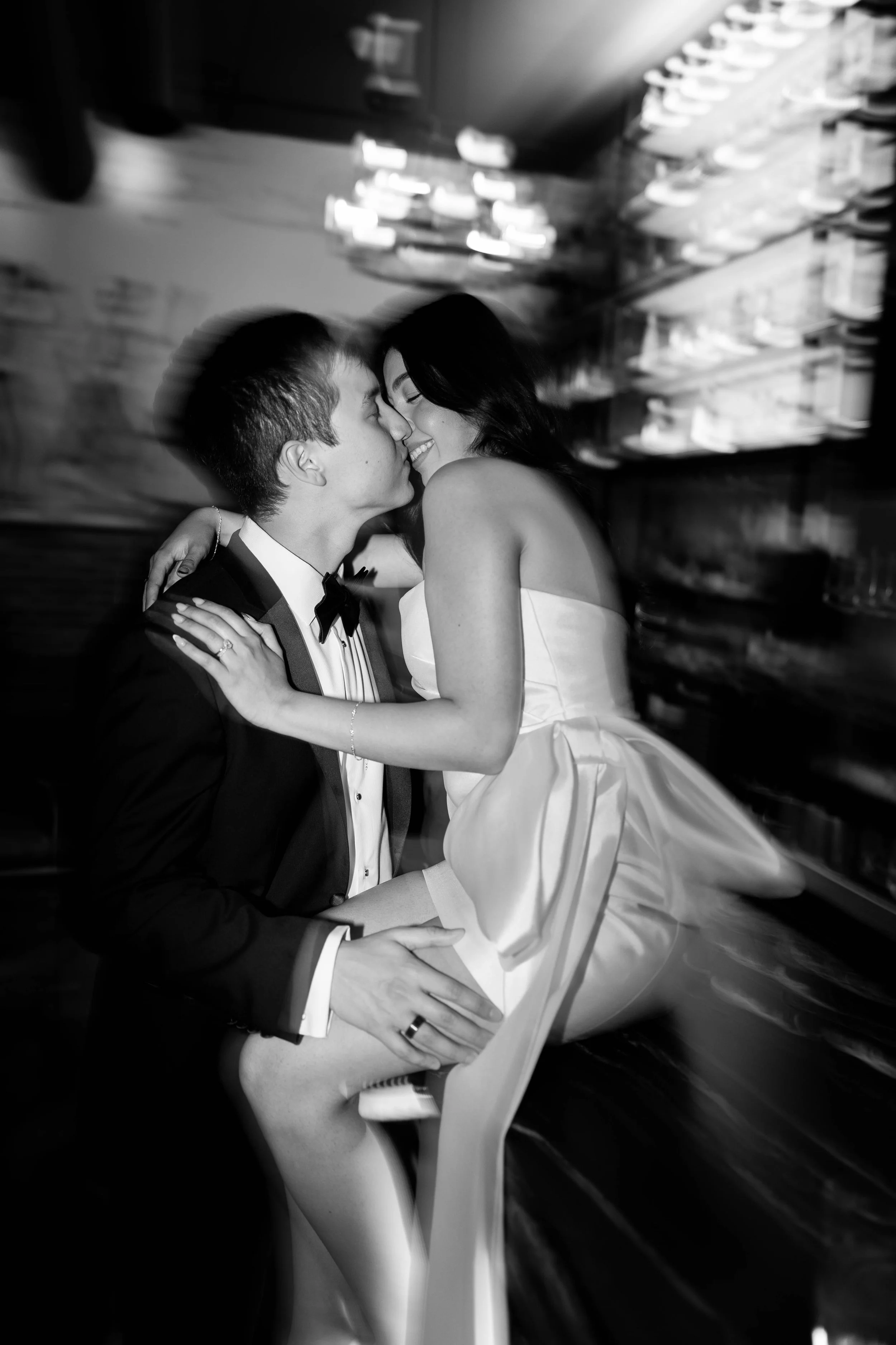 A black and white photo of a bride and groom sharing a kiss and smiling, with the bride sitting on the groom's lap inside a bar or restaurant. The groom is dressed in a tuxedo and bowtie, and the bride is wearing a wedding dress.