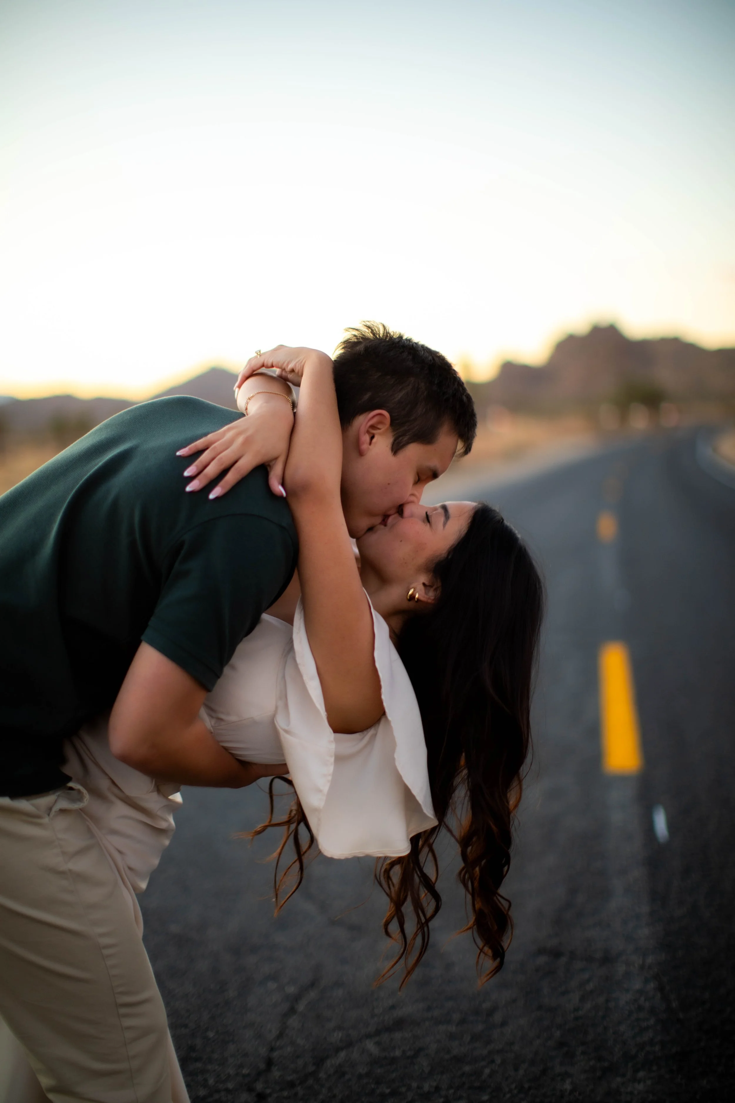 A couple sharing a kiss on a deserted road at sunset, with mountains in the background.