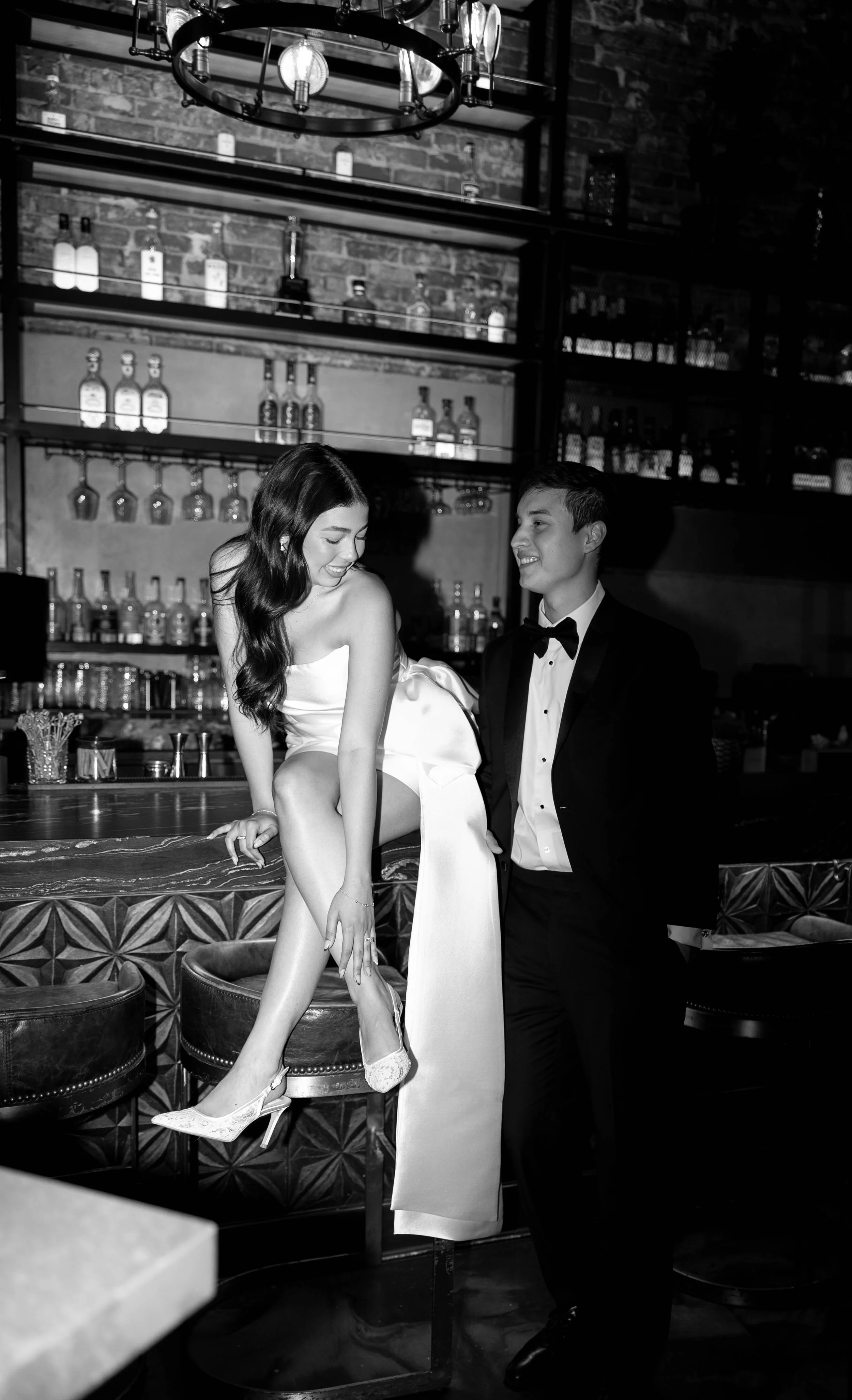 A black and white photo of a woman in a strapless dress sitting on a bar counter with her legs crossed and a man in a tuxedo standing next to her, smiling at her in a bar or lounge setting.