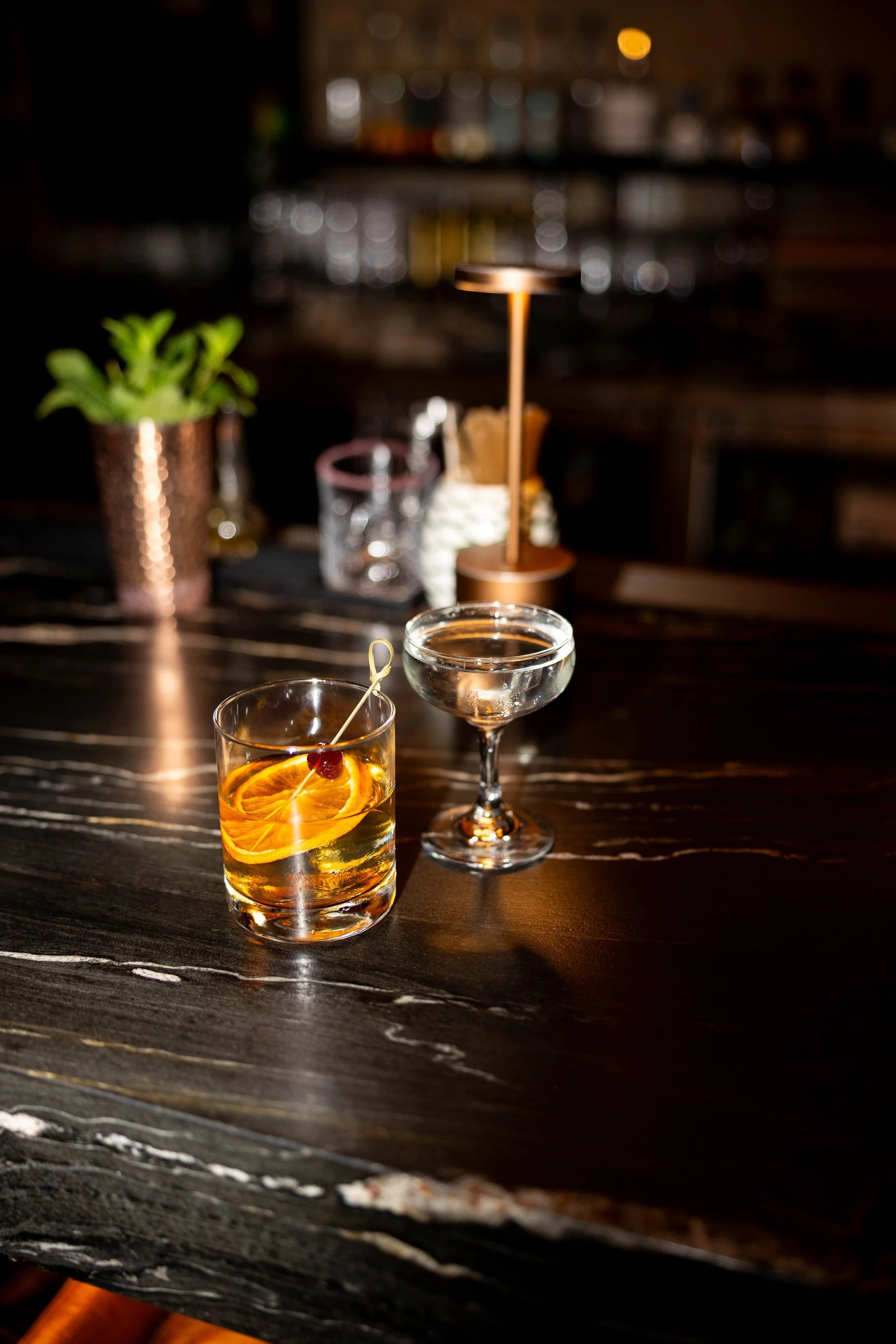A bar counter with two cocktails, one garnished with orange slices and a cherry, and the other in a coupe glass, with bar supplies and a plant in the background.