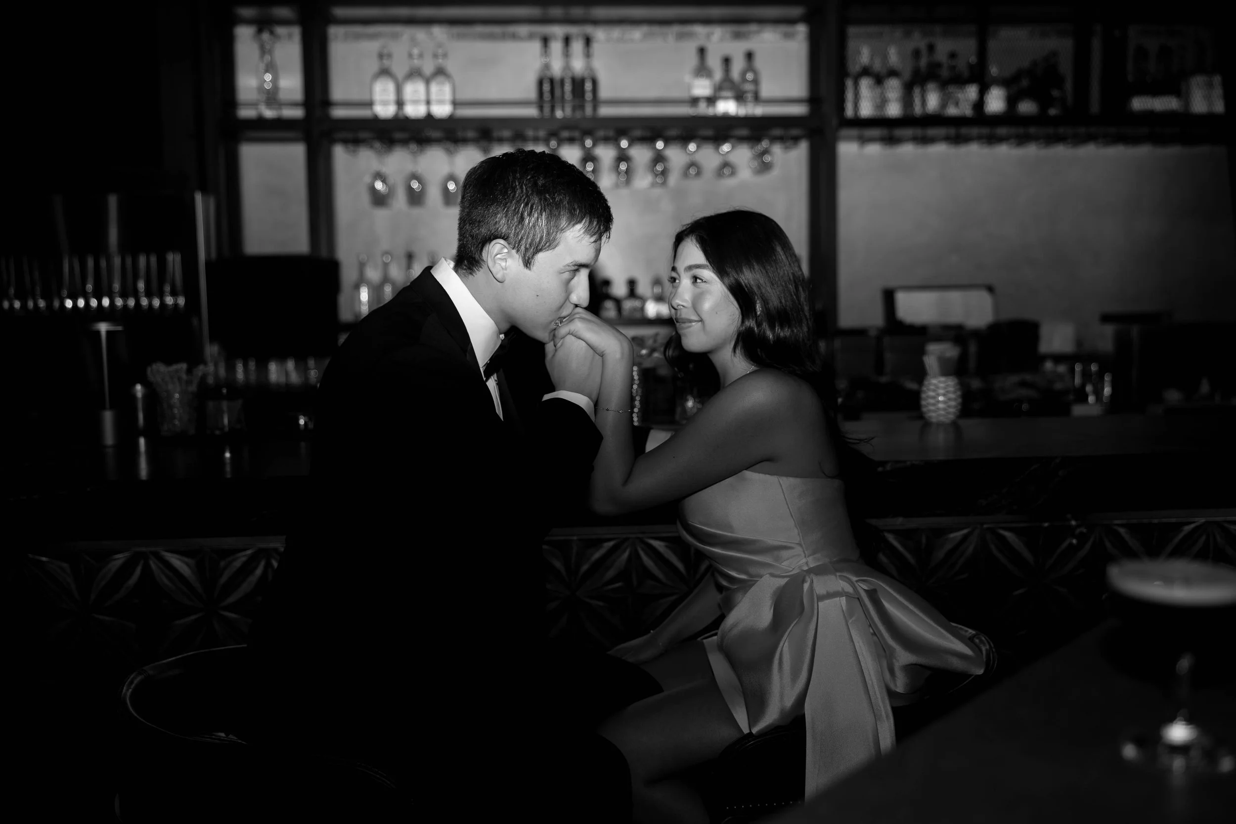 A young man and woman sitting close together at a bar, with the man leaning in and the woman smiling, in a dimly lit setting.