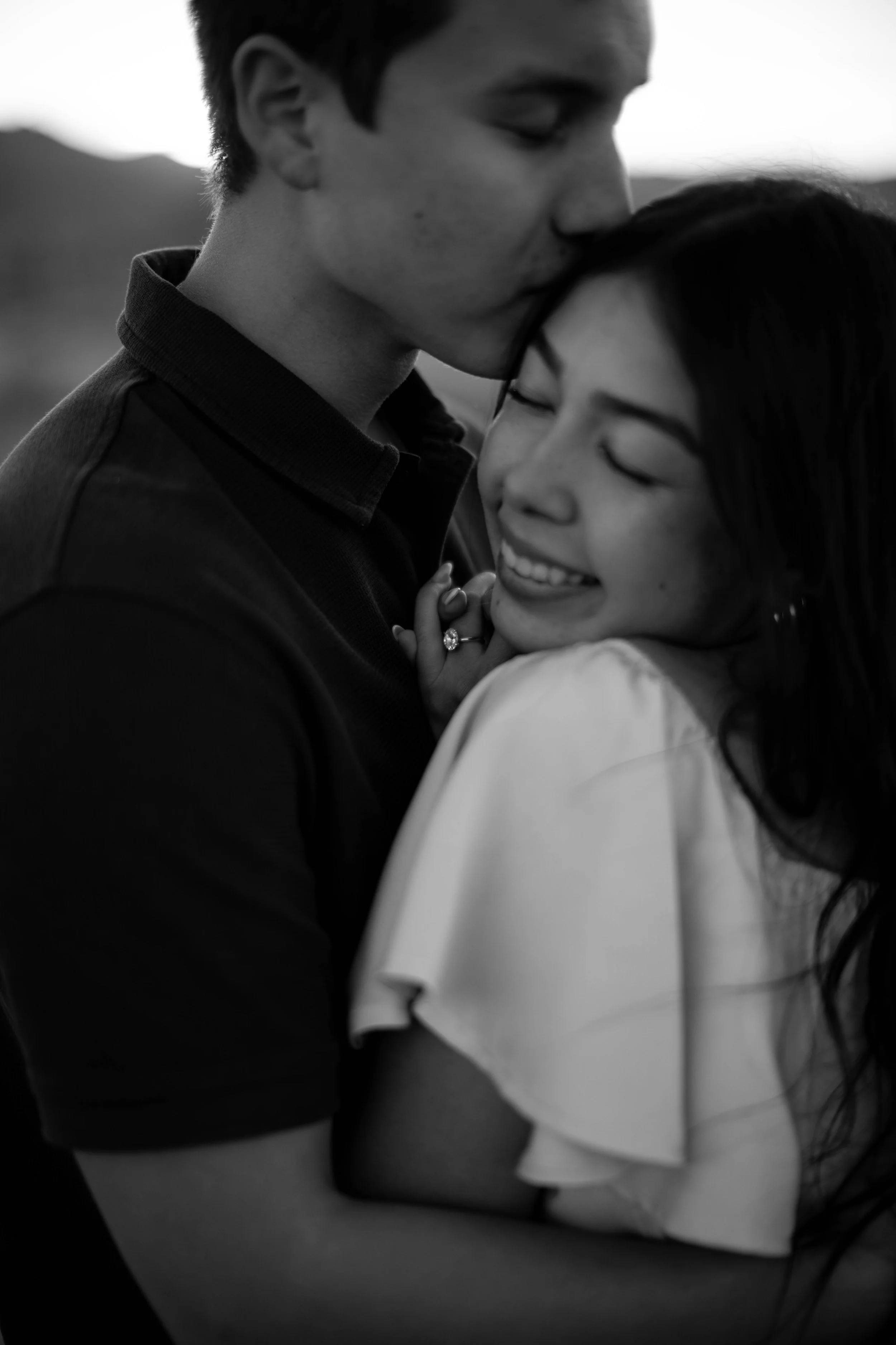 A black and white photo of a couple embracing, with the man tenderly kissing the woman's forehead. The woman is smiling and her eyes are closed.