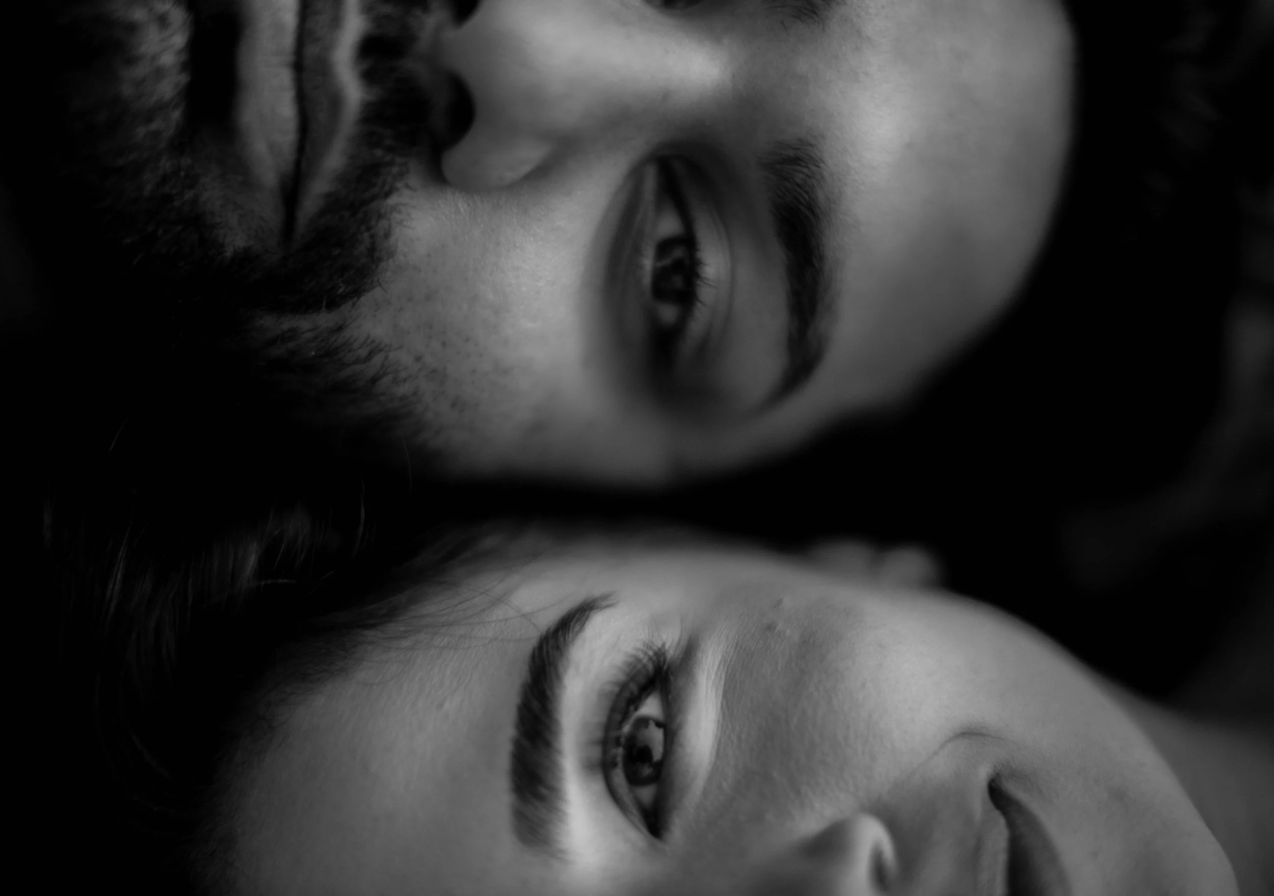 Close-up of a man with a beard and a woman with long hair, both lying down, facing each other with their eyes open, in black and white.