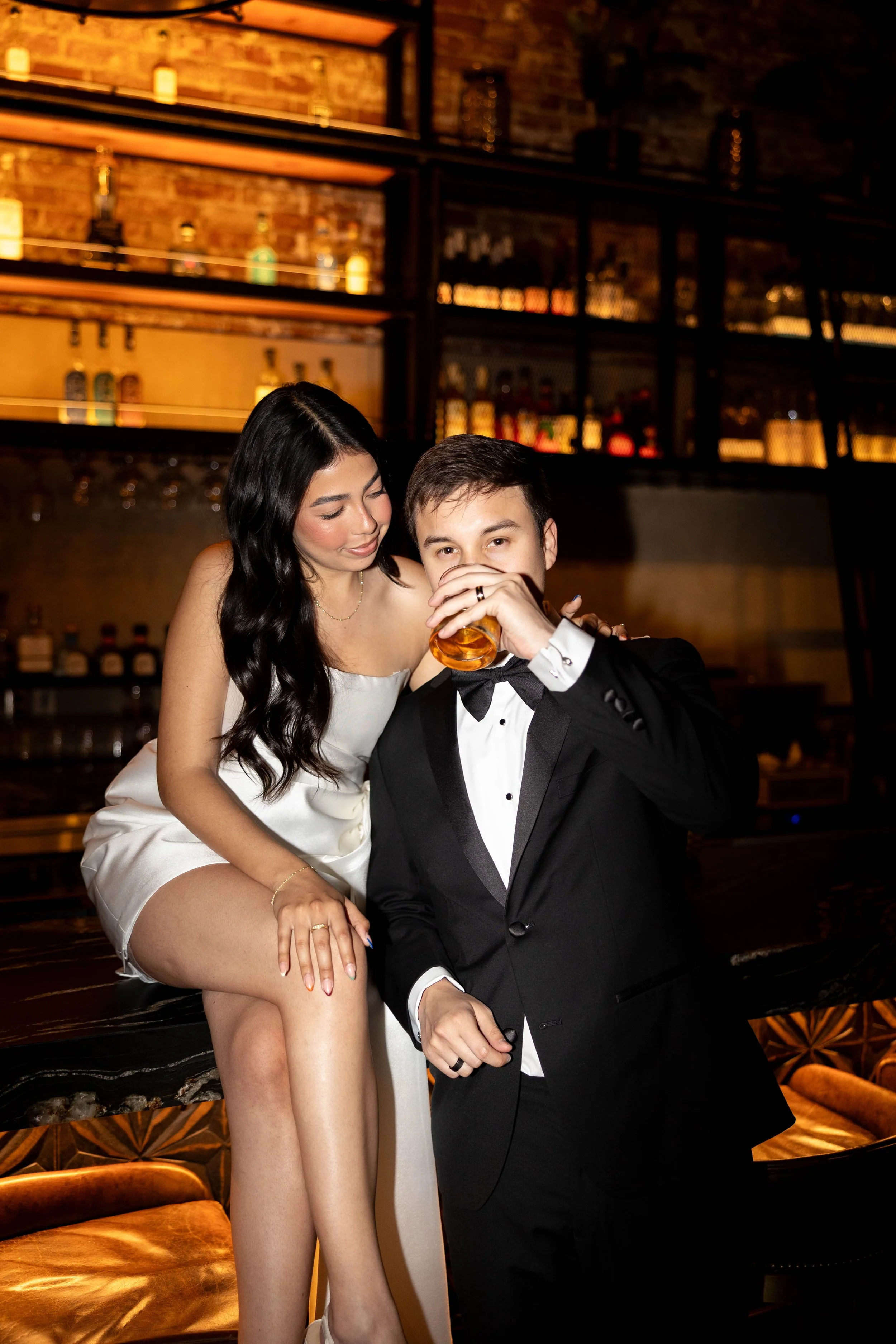 A woman in a white dress with long black hair and a man in a black tuxedo with a bowtie are at a bar. The woman is sitting on the bar with her legs crossed, looking at the man, who is drinking from a glass of whiskey.