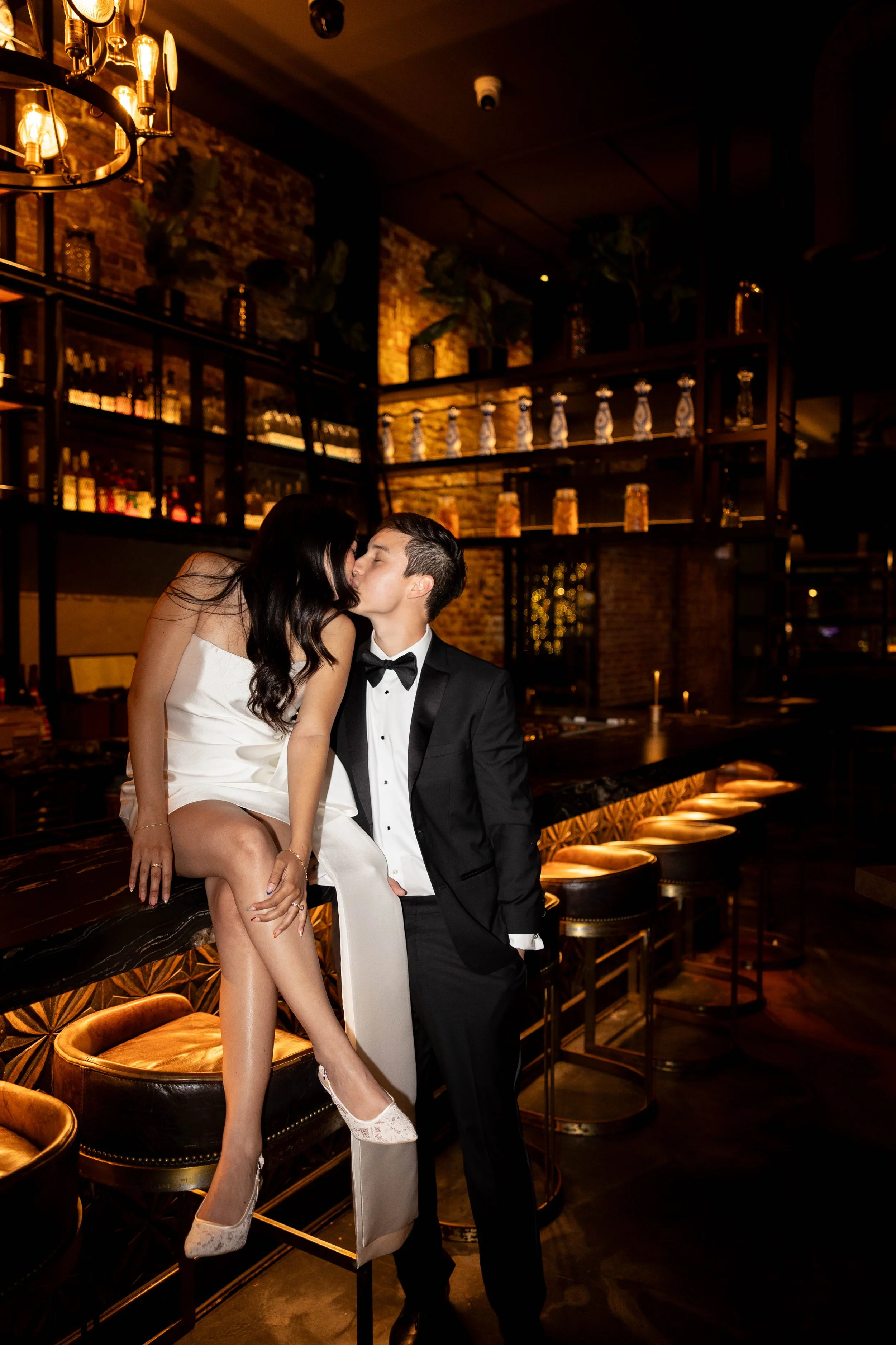 A woman in a white dress and a man in a black tuxedo sharing a kiss in a dimly lit bar or lounge.
