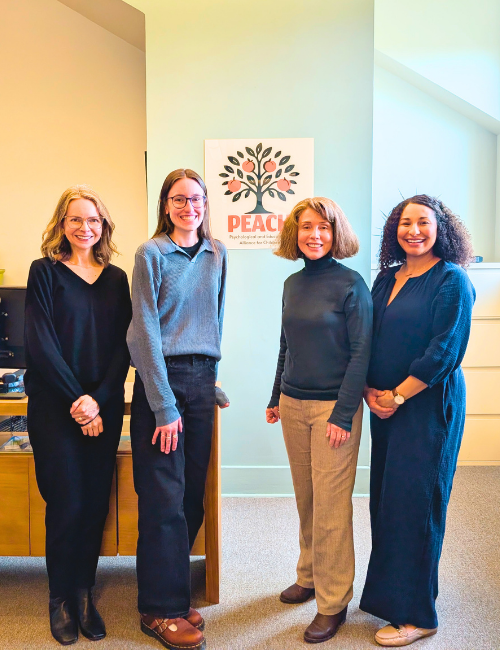 Four psychologists standing in a psychoeducational testing center, smiling at the camera. Behind them is a sign with the word "PEACH".