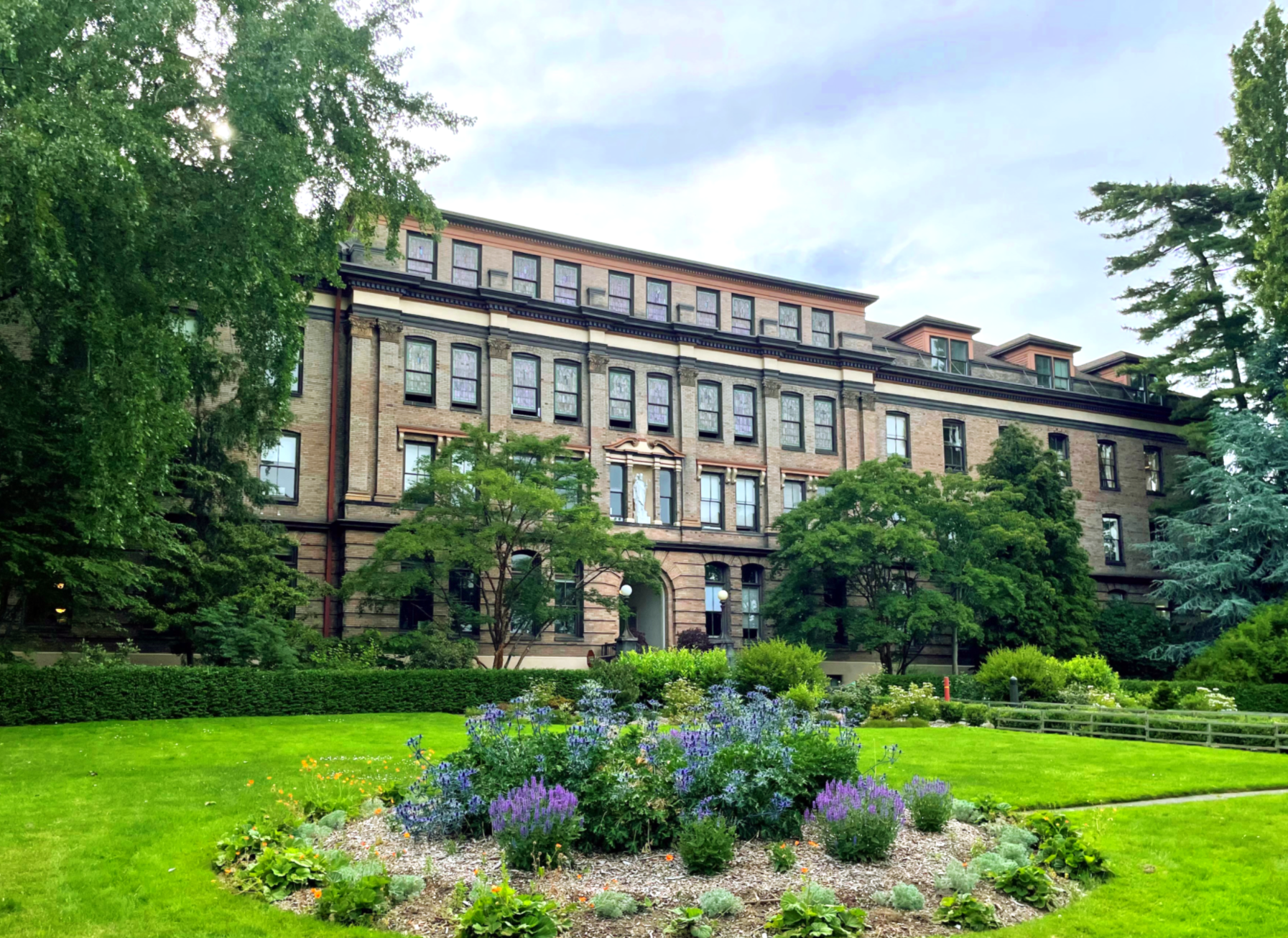 A large, historic, multi-story brick building behind a lush green garden with various trees and colorful flowering plants housing a school, an events location, and several psychological services.