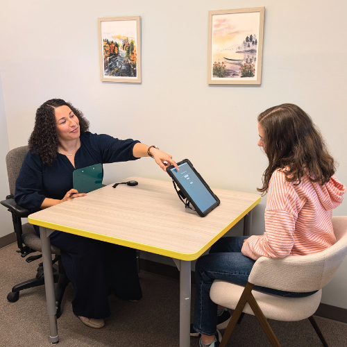 A psychologist with dark curly hair and a navy blue shirt is testing a young girl with long brown hair, a pink striped hoodie, and jeans, for ADHD.