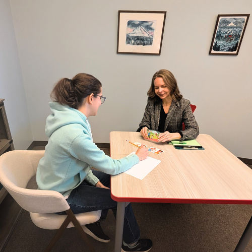 A child psychologist and a teen girl are sitting across from each other at a wooden table in a therapy room. The teen girl is writing her responses to an IQ test on paper, and the psychologist is smiling, holding a test .