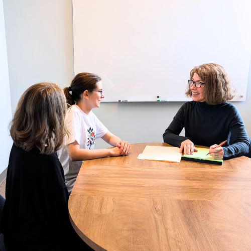 A child psychologist is sitting at a conference table in a meeting room, explaining psychoeducational test results to an adolescent girl and her mother.