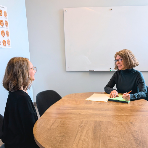 Two women sitting across from each other at a wooden table in a bright room, with a whiteboard on the wall behind them, engaged in a conversation or interview.