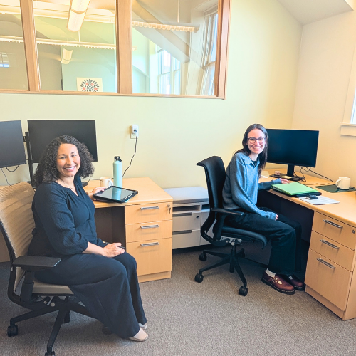 Two psychologists sitting at desks in an office to score academic tests. They are smiling at the camera. One woman has curly hair, wearing a dark top, and the other has straight hair, glasses, a blue sweater, and dark pants.