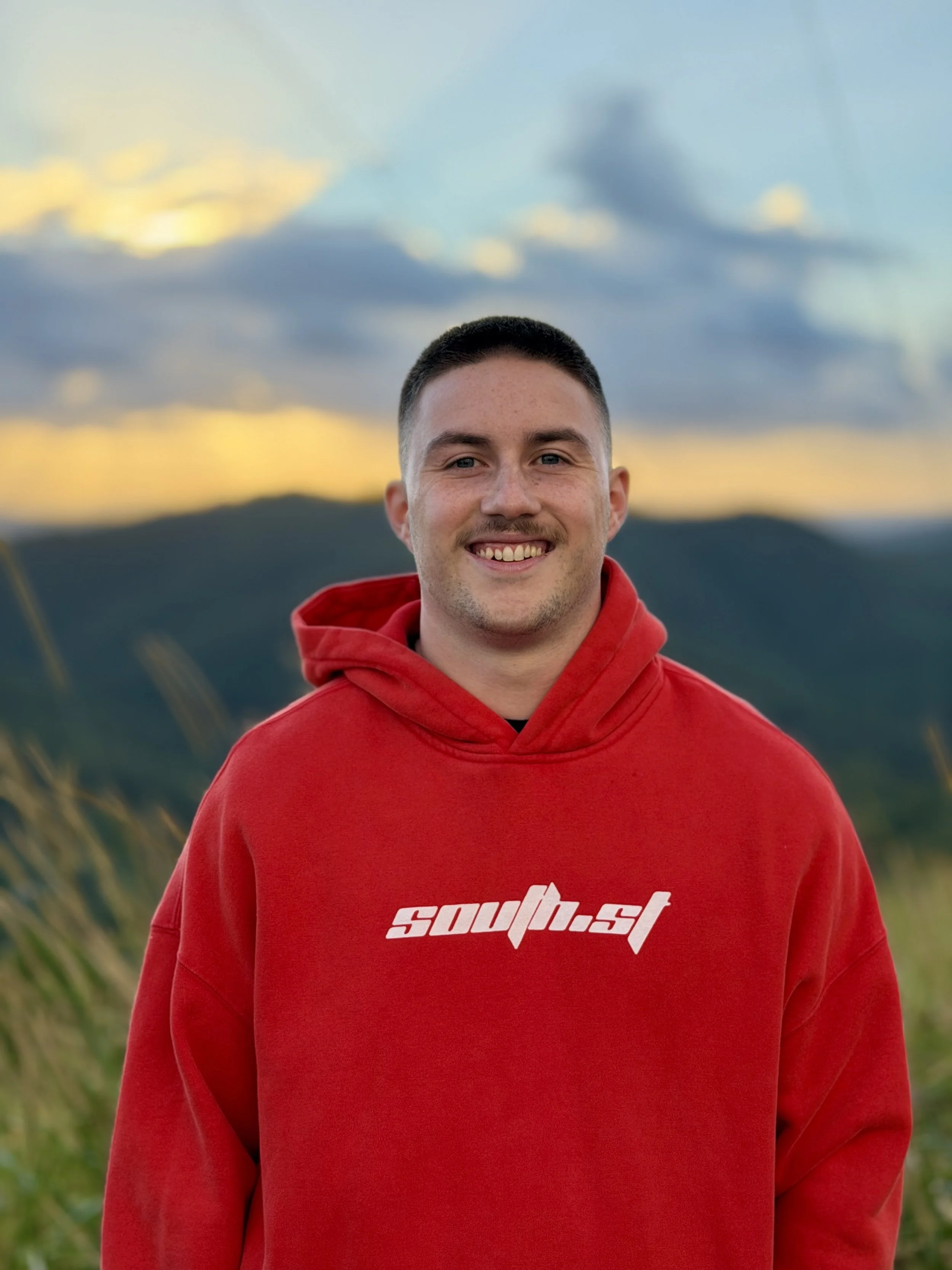 A smiling young man with short dark hair, wearing a red hoodie with white text, standing outdoors with mountains and a cloudy sky in the background.