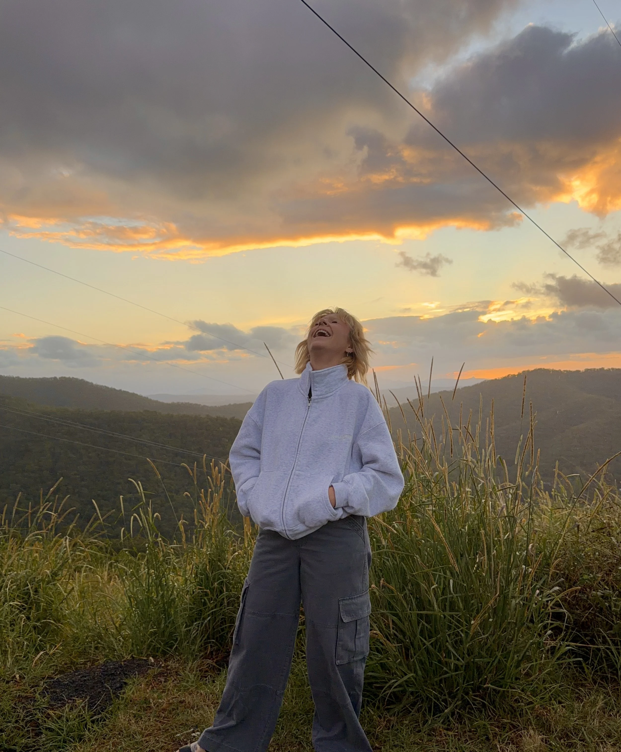 Person with blonde hair, wearing a white jacket and dark pants, standing outdoors in a field of tall grass during sunset, with a dramatic sky filled with dark and orange clouds.