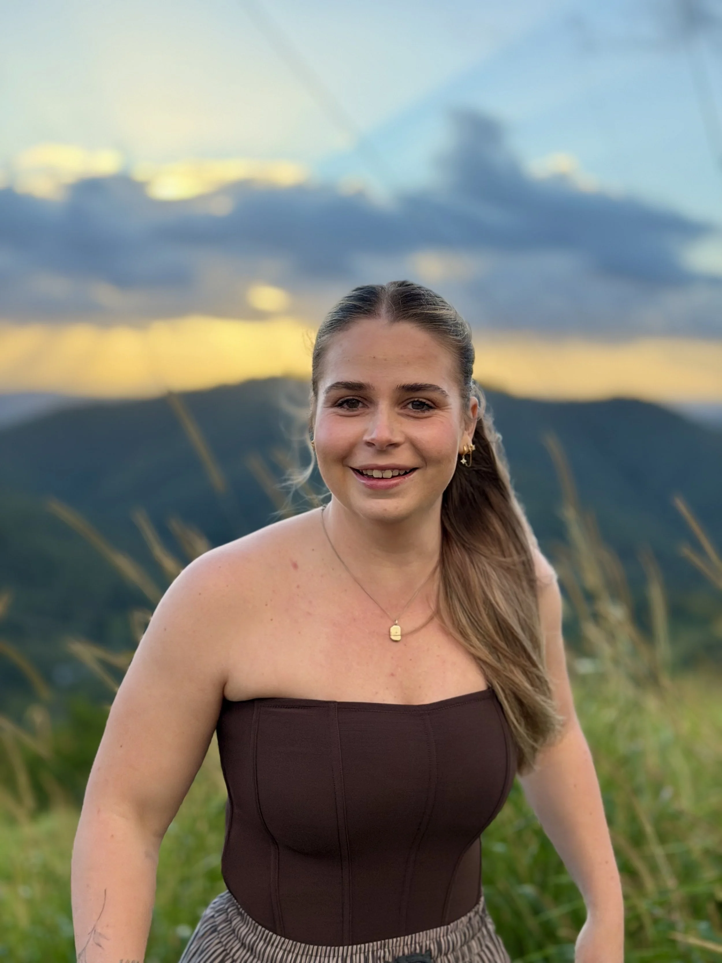 A woman smiling outdoors with mountains in the background during sunset, wearing a strapless brown top, earrings, a necklace, and patterned pants.