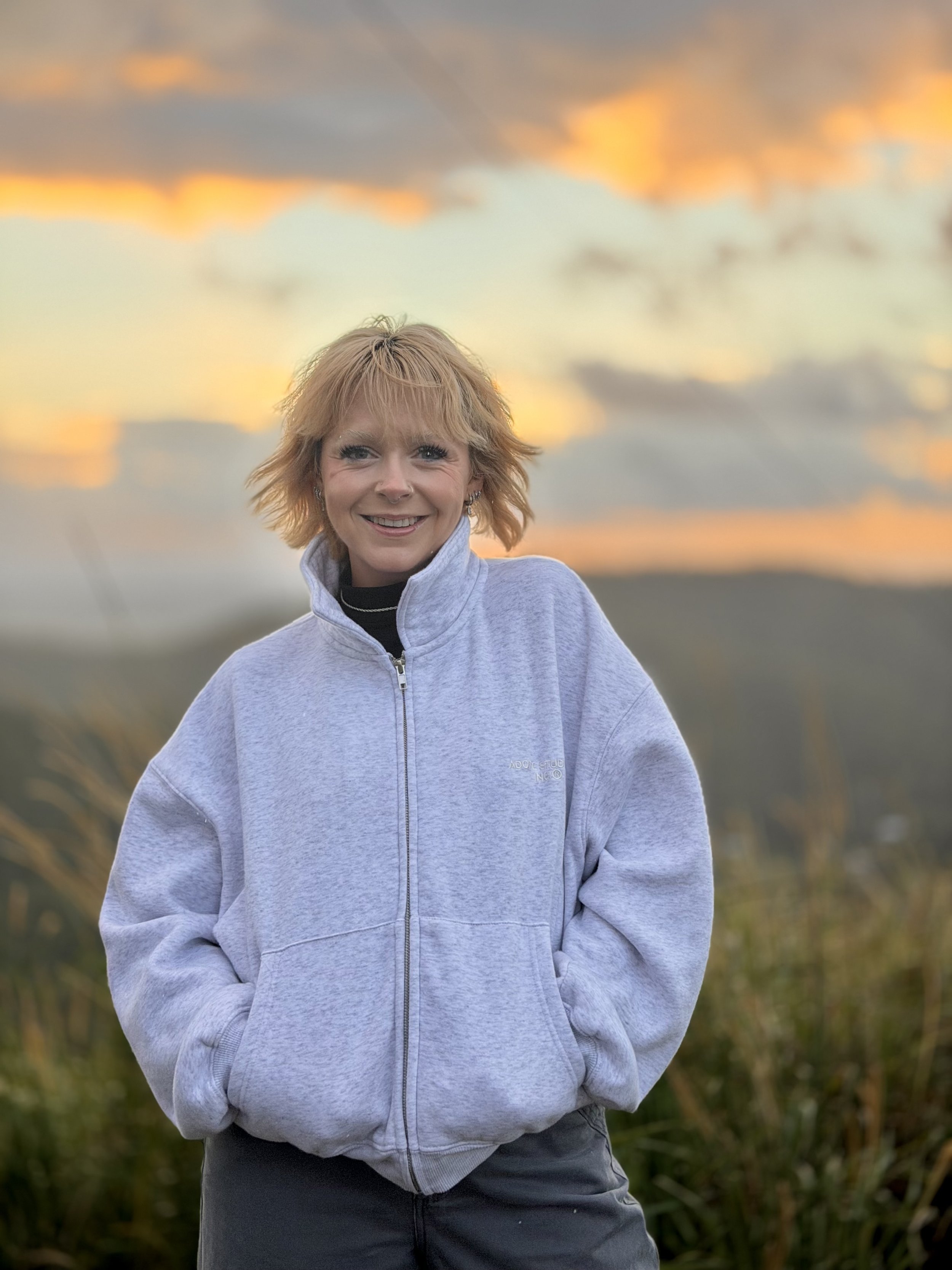 A woman with short blonde hair smiling outdoors at sunset, wearing a light gray zip-up sweatshirt and dark pants, standing in a grassy field.