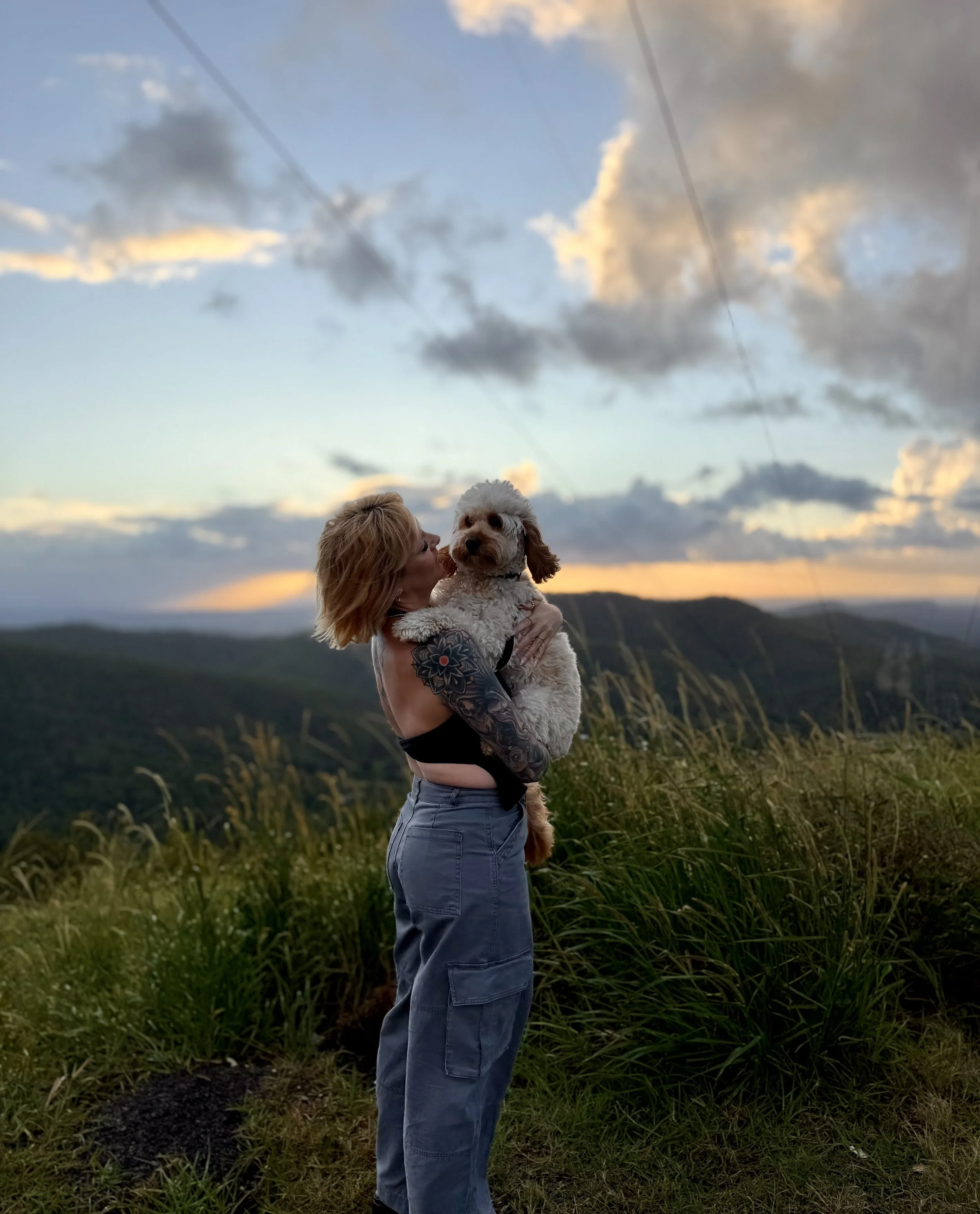 A woman with tattoos on her arm holding a fluffy white dog during sunset outdoors, with tall grass in the foreground and a sky with clouds in the background.