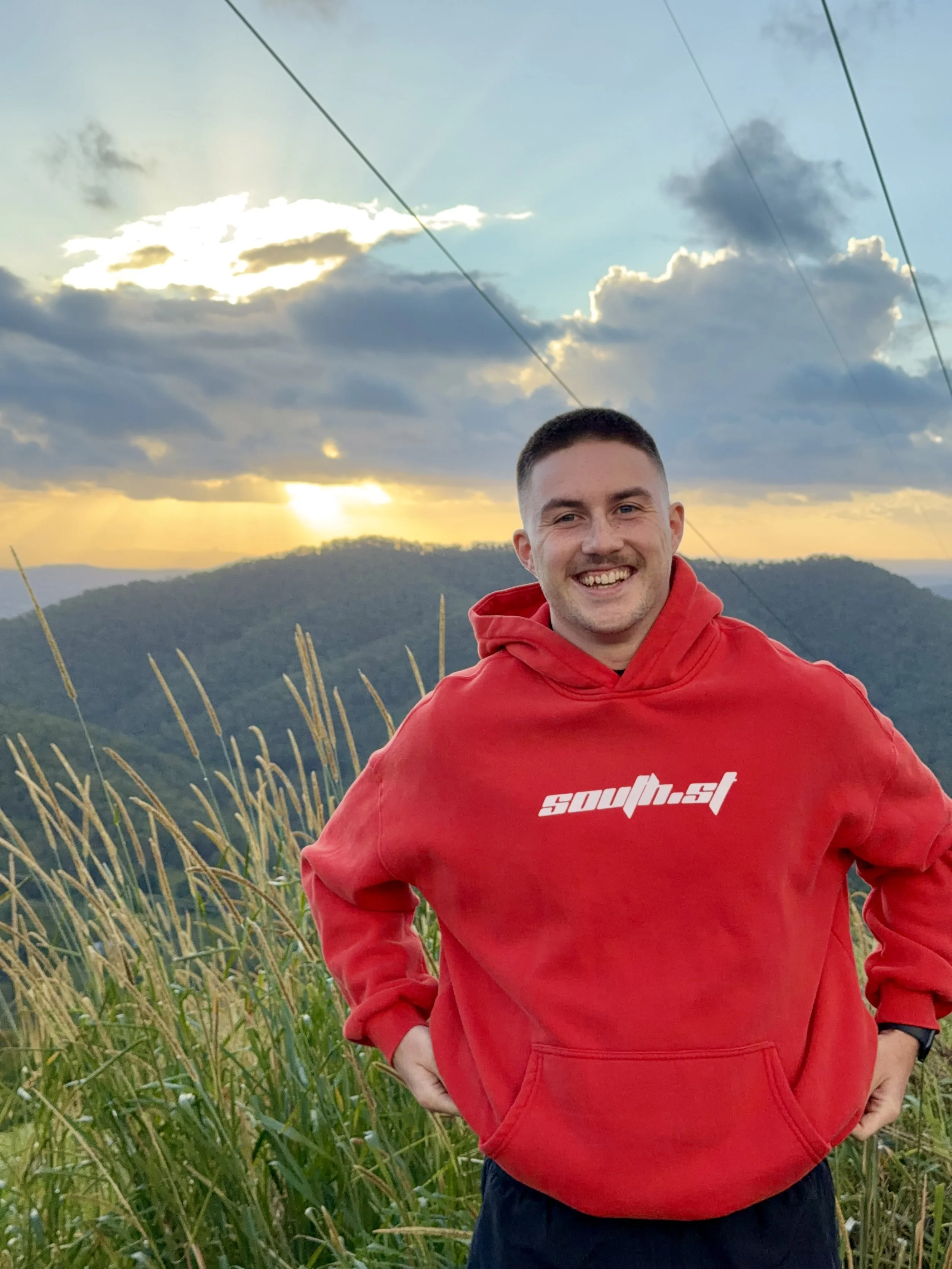 A young man smiling, wearing a red hoodie with white text, standing outdoors during sunset with cloudy skies and a mountain in the background.
