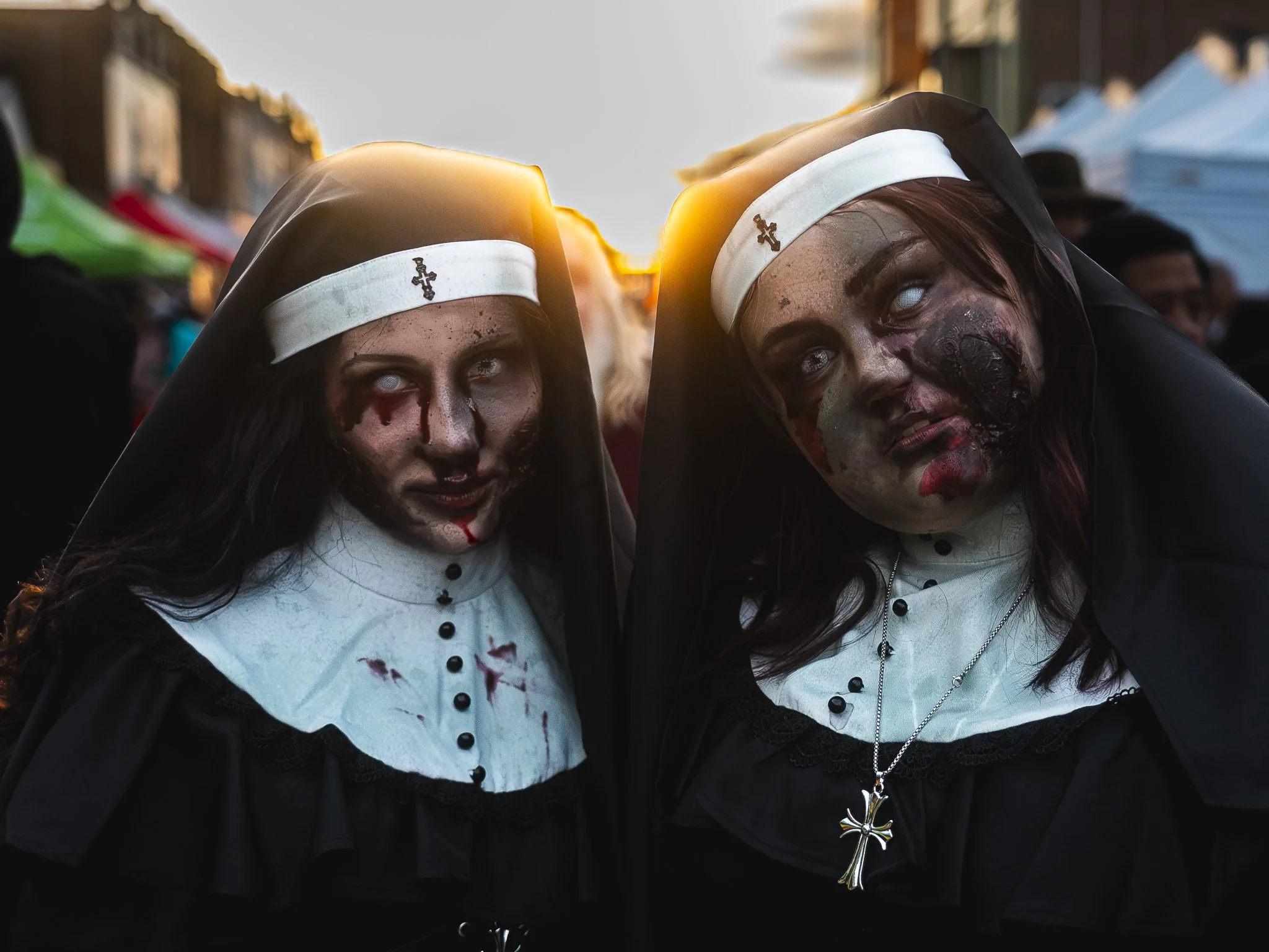 Two women dressed as nuns with zombie makeup, including fake blood and Pale eyes, standing outdoors at sunset with tents in the background.