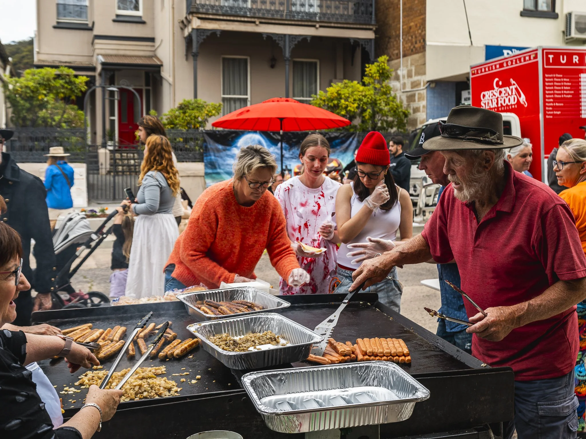 People grilling and serving hot dogs at an outdoor event, with a red umbrella overhead and a food truck in the background.