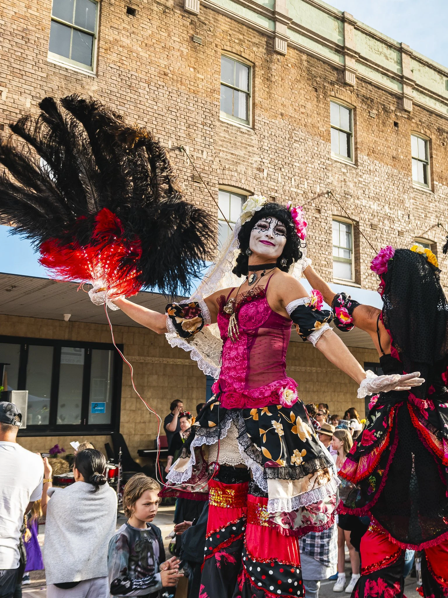 Person dressed in colorful costume with face paint, holding large black and red feathered fan, participating in a street parade or festival.