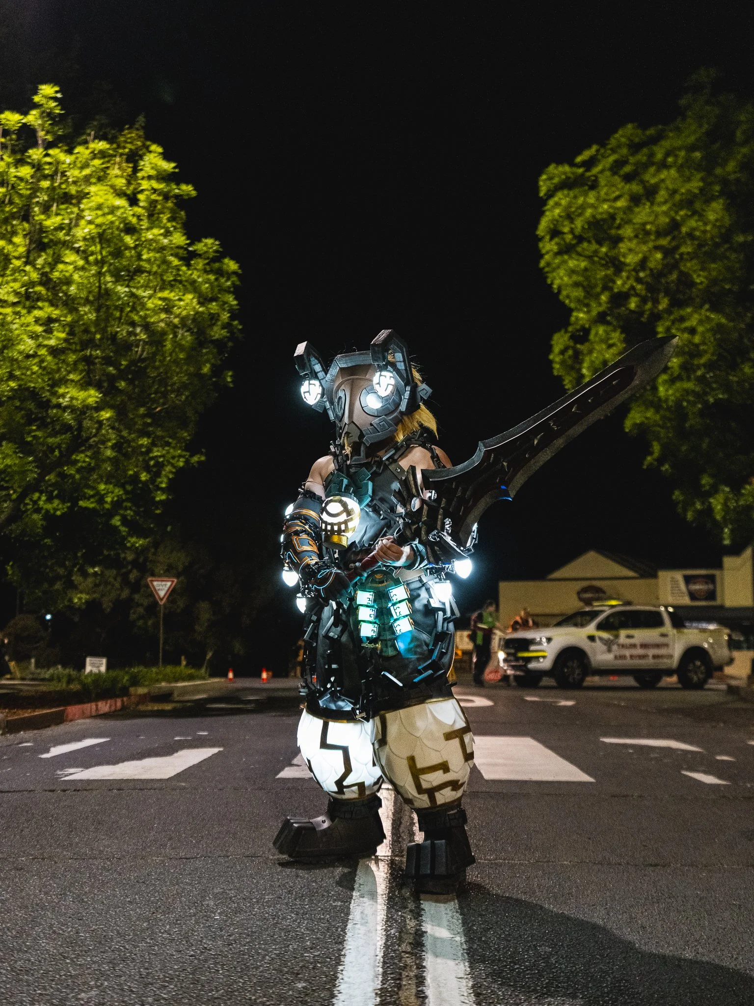 Person in a futuristic armor costume with armor helmet, holding a large sword, standing in the middle of a street at night with trees and parked cars in the background.