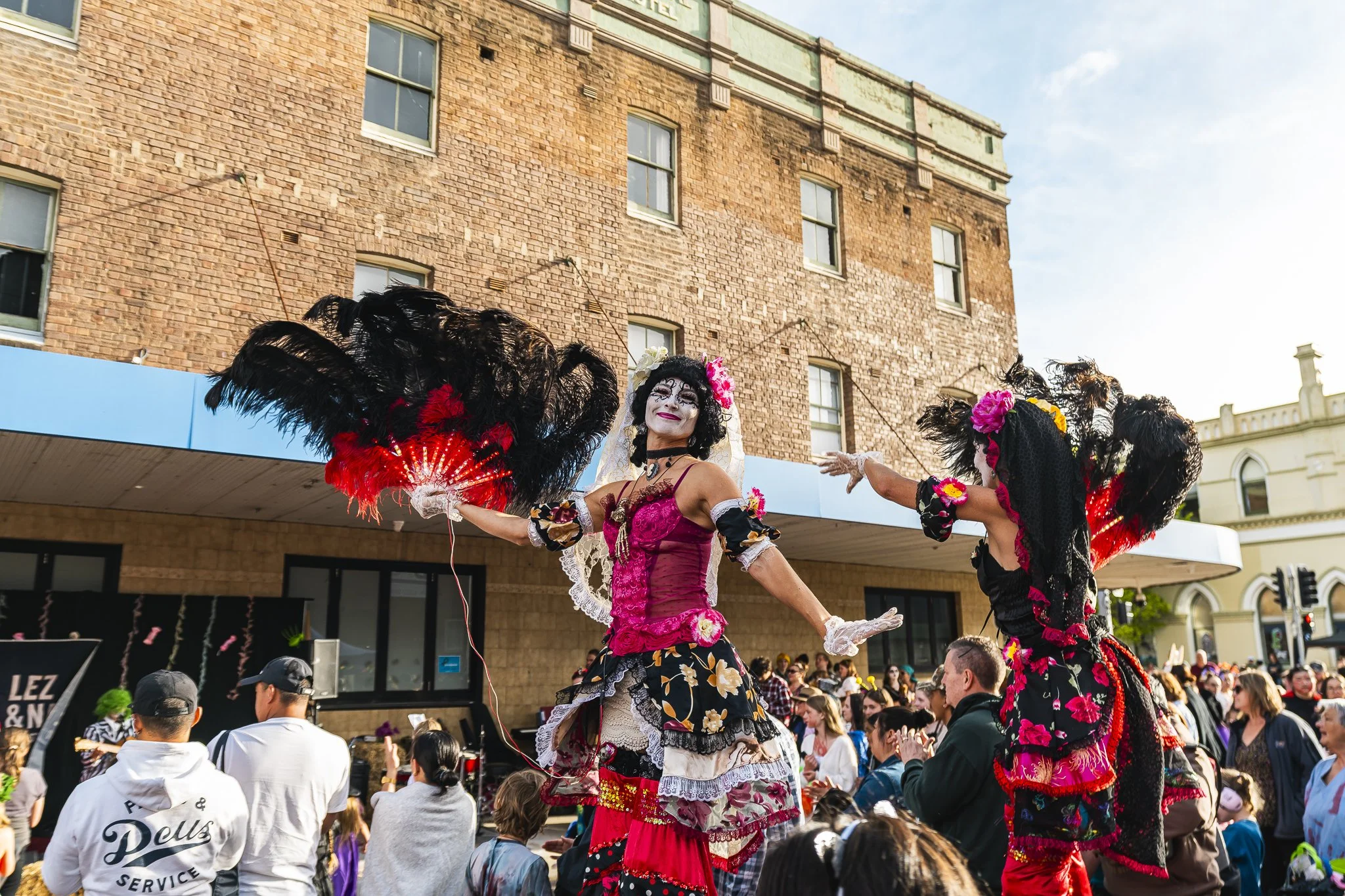 Street performers in colorful costumes dancing at a festival, with a crowd watching in front of brick and historic buildings.