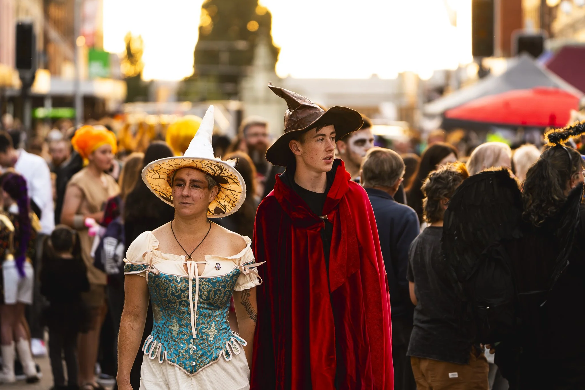 People dressed in costumes at a street event or festival, with two individuals in fantasy or witch costumes in the foreground and a crowd behind them.