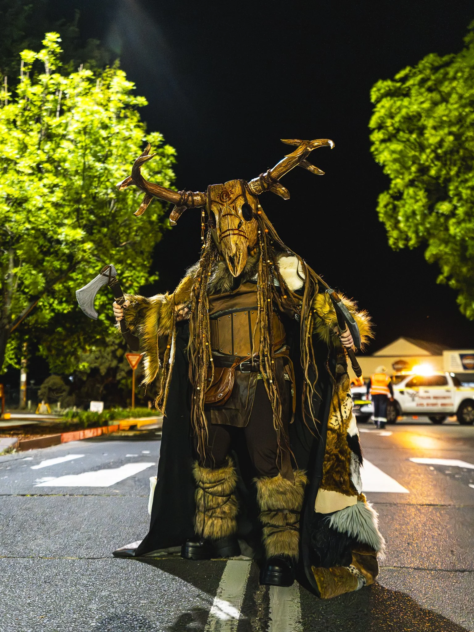 A person dressed in a detailed costume with a large wooden mask and antlers, holding an axe, standing on a street at night surrounded by trees and vehicles.
