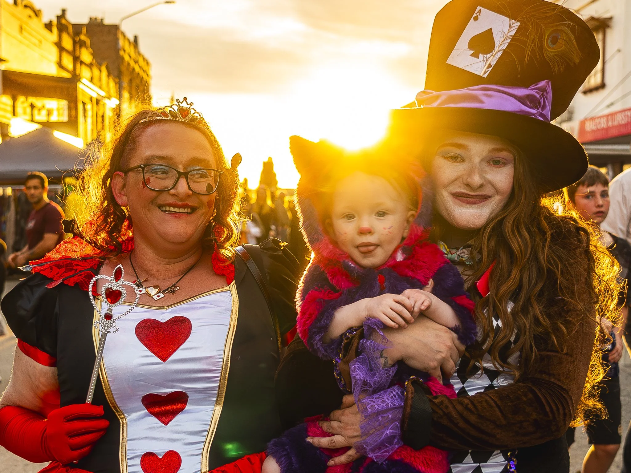 Two women dressed in costumes, one as the Queen of Hearts and the other as a witch, holding a young girl in a red and purple costume, at a street event during sunset.