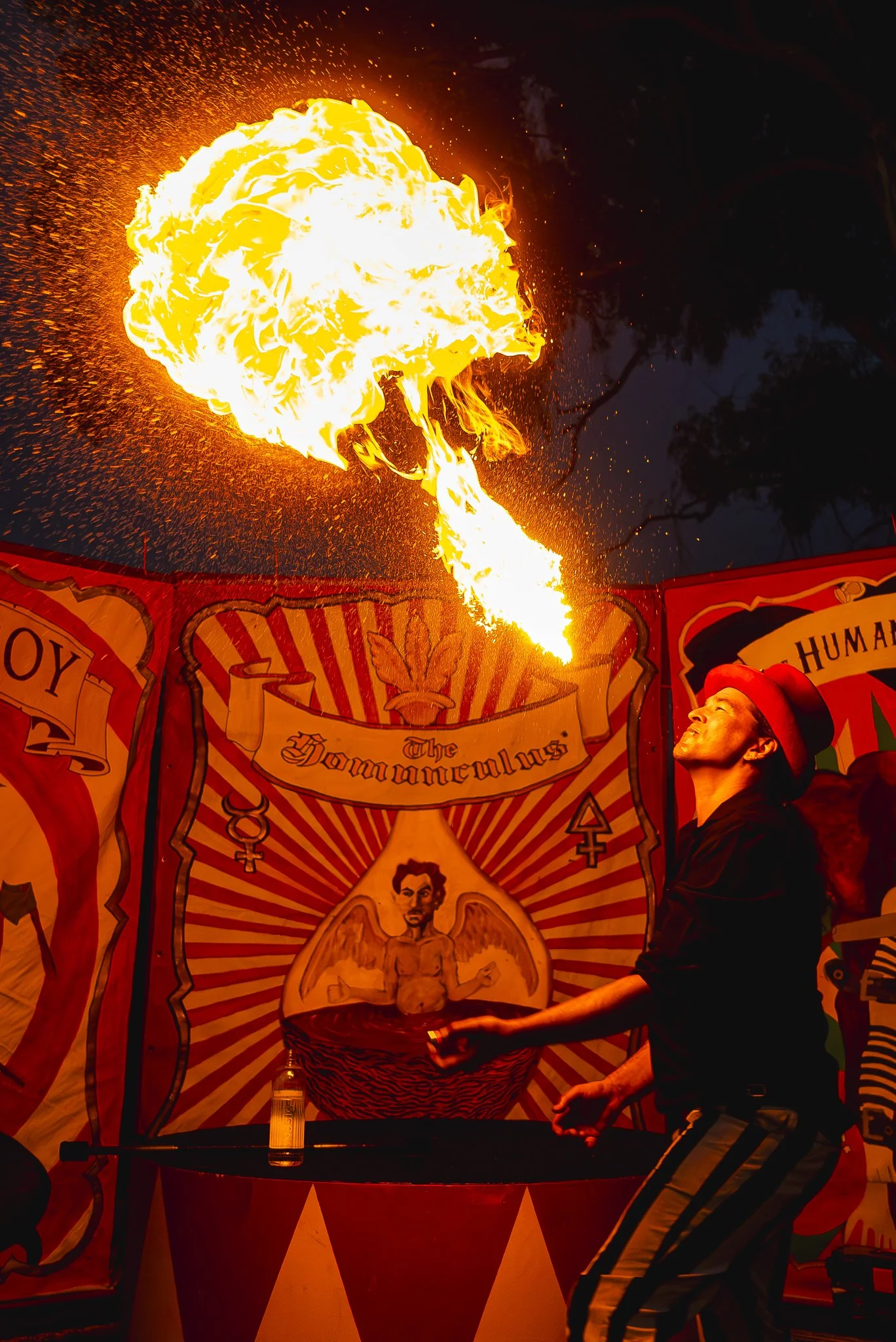 A performer in red hat, black shirt, and striped pants creates a fireball during a nighttime circus or street performance, with colorful banners in the background.